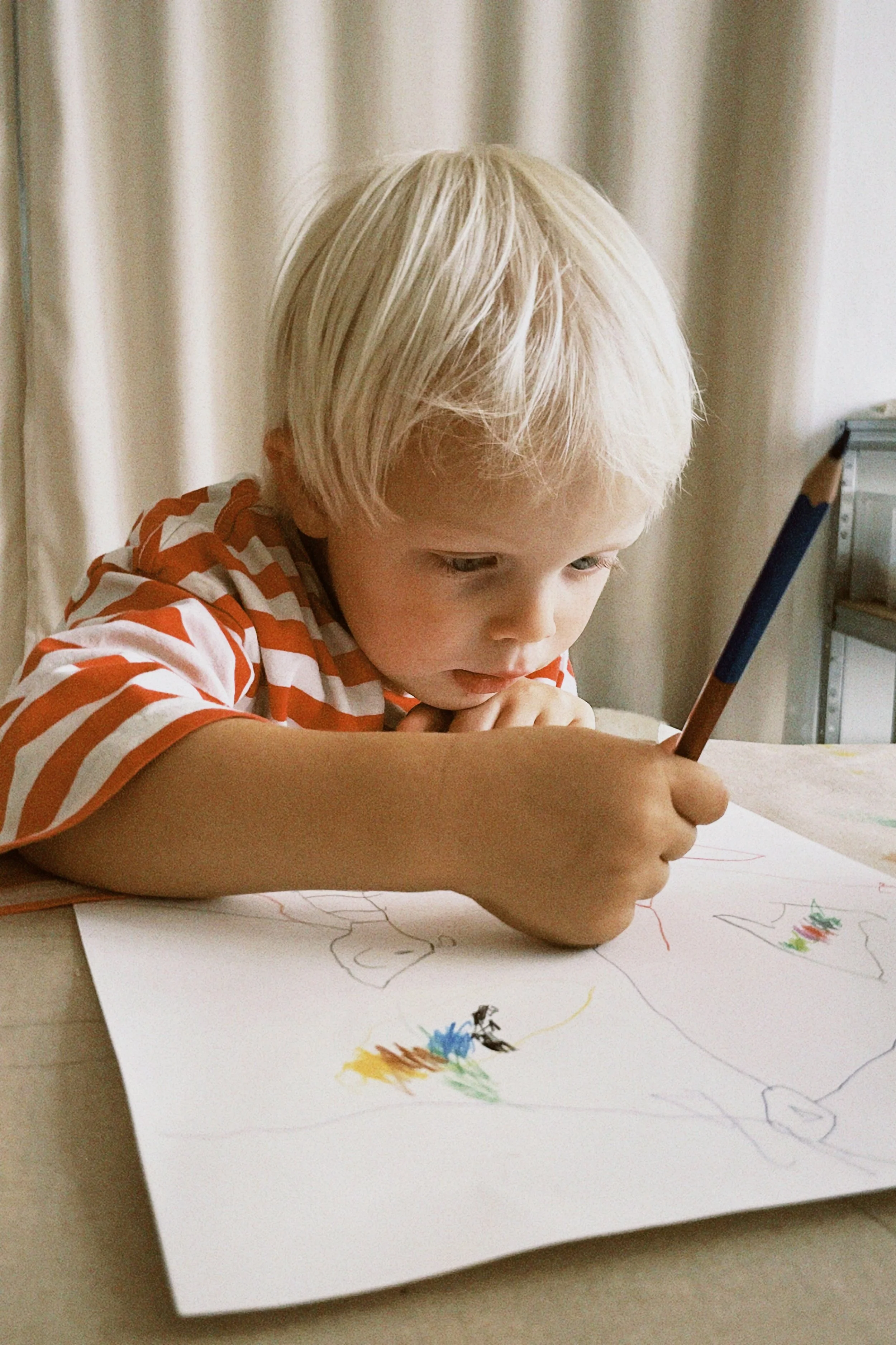 Ein Junge mit blondem Haar zeichnet mit Buntstiften auf Papier, das auf einem Tisch liegt.