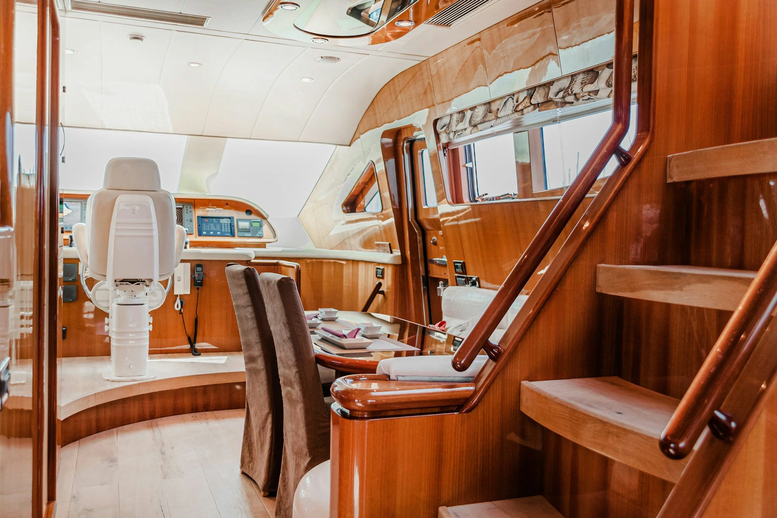 The interior of a yacht showcasing a control area with a white captain's chair, wooden cabinetry, window blinds, and a dining table with chairs.