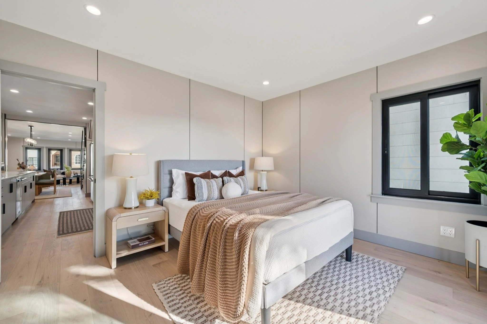 A modern bedroom with a bed featuring white and brown pillows, beige blanket, matching bedside tables with lamps, a window with black frame, and light-colored wooden flooring.