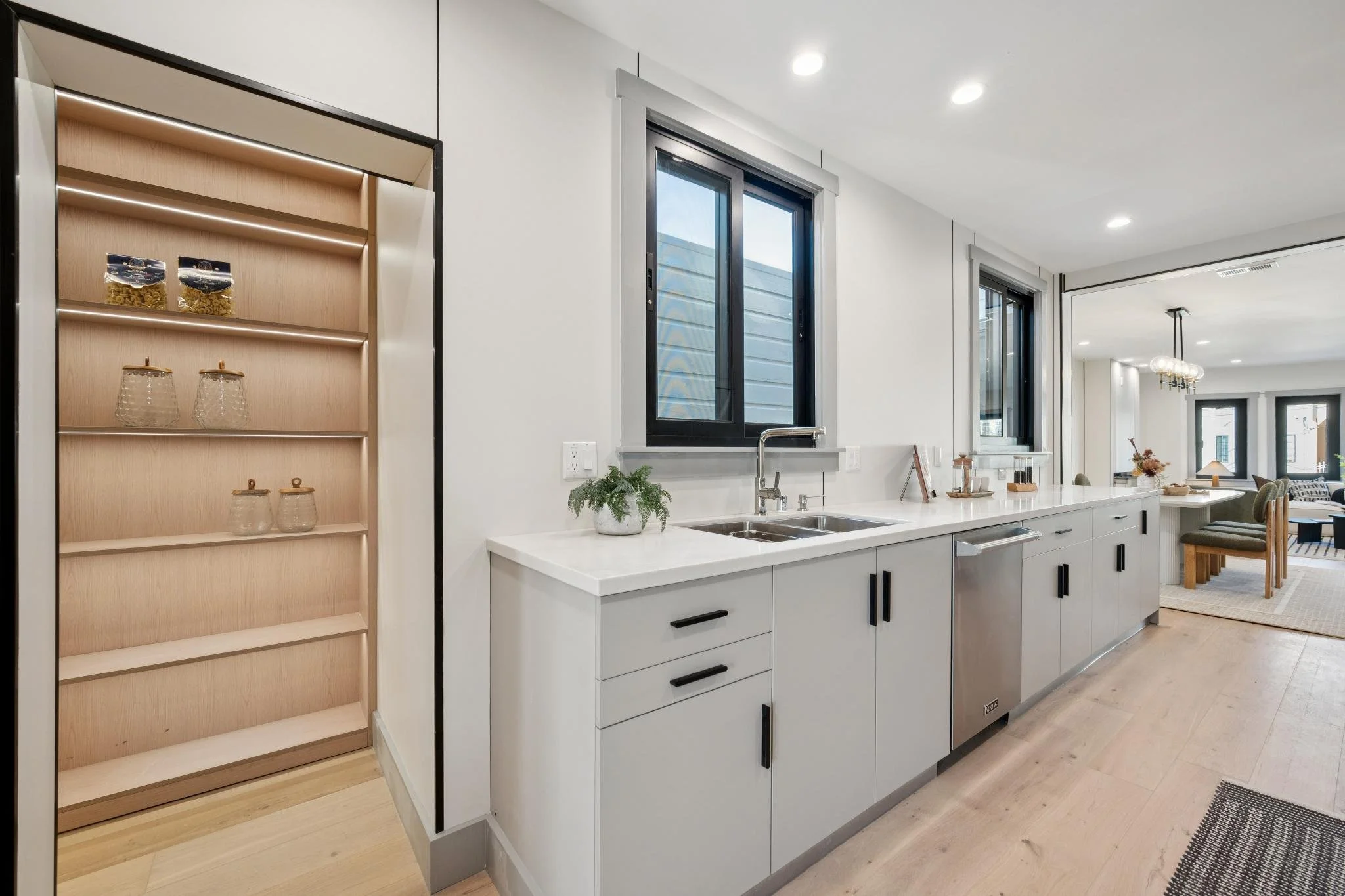 Modern kitchen with white cabinetry, black handles, and a stainless steel dishwasher. Two black-framed windows above the sink, with a small potted plant and decorative items on the countertop. An empty wooden shelving unit on the left. Open layout wi