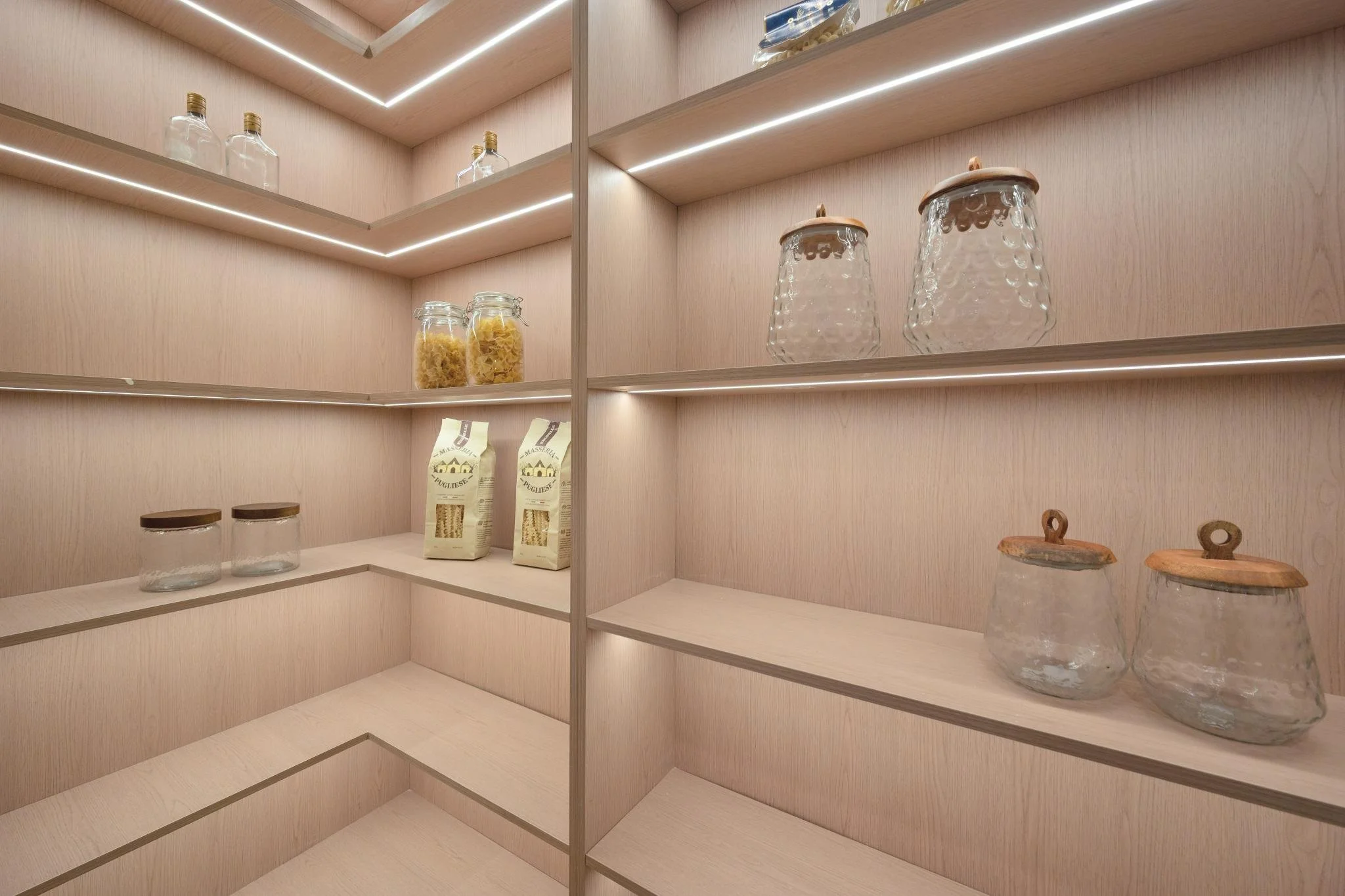 Empty wooden shelves in a modern kitchen cabinet with glass jars, pasta, and other kitchen items, illuminated by LED strip lighting.