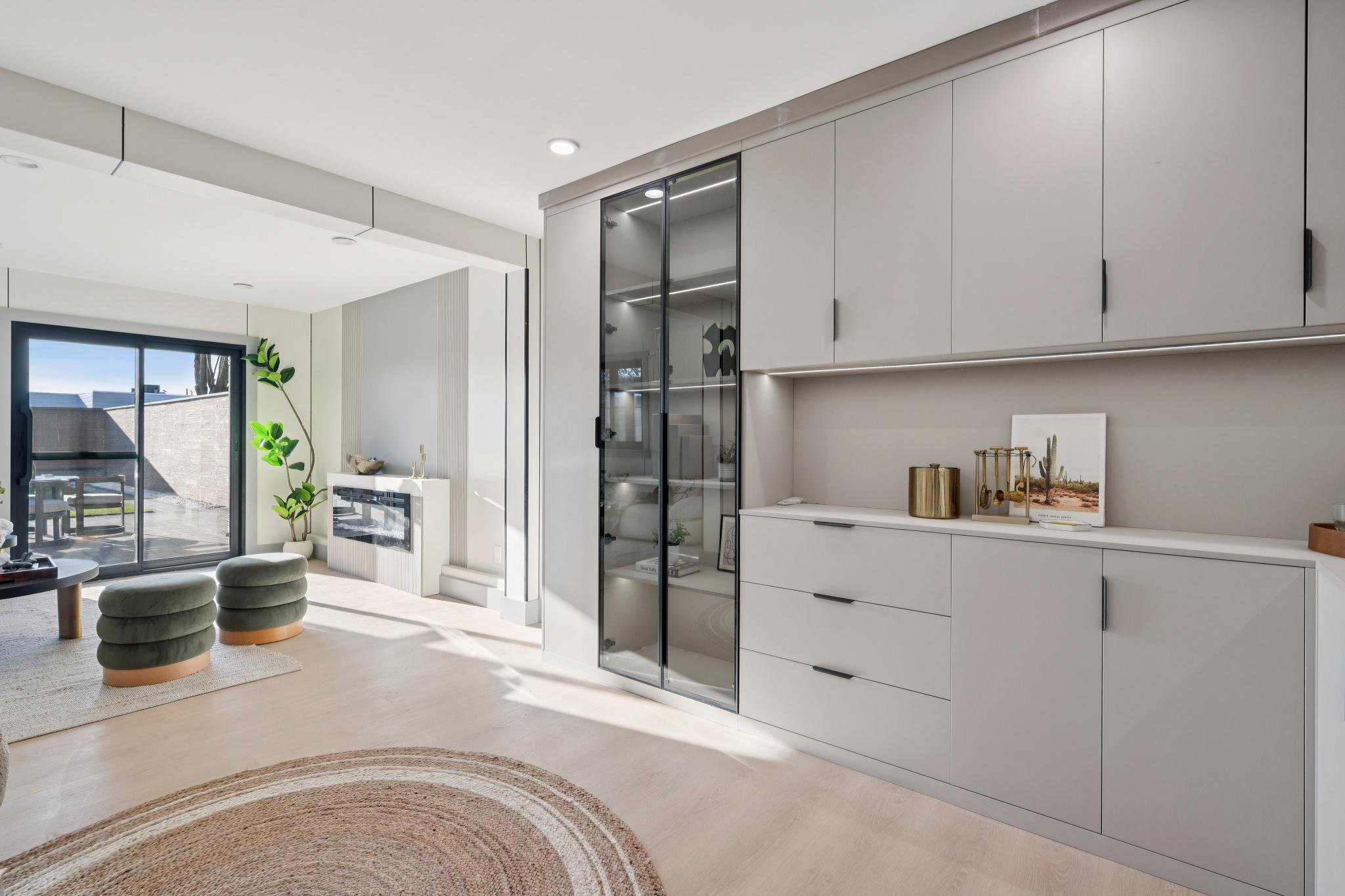 Modern living room with white walls, light wood flooring, and large glass sliding door leading outside. Green plant by the window, circular rug, and minimalist furniture include two green ottomans and a light-colored sideboard with decorative items.