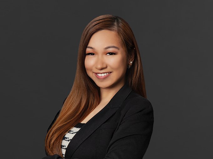 Professional portrait of a young woman with long brown hair, wearing a black blazer and striped top, smiling against a gray background.