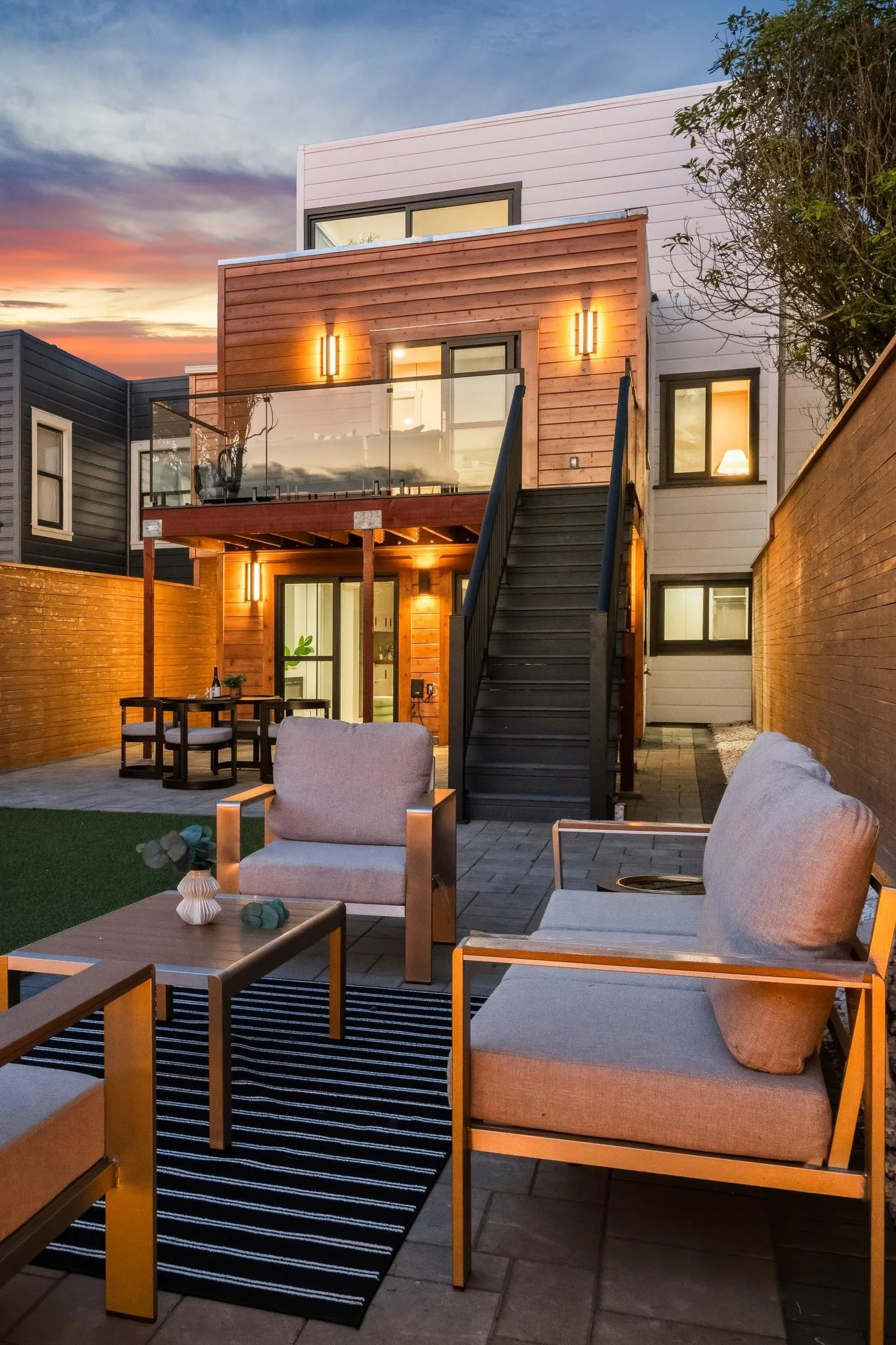 Modern two-story house with an outdoor patio at sunset, featuring seating area and a staircase leading to the second floor balcony.