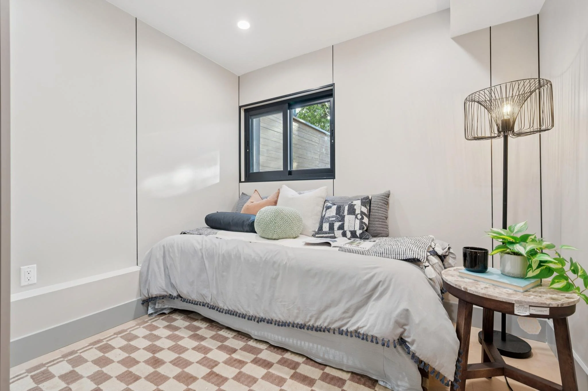 A cozy bedroom with a white bed, a plaid blanket, cushions, a side table with a plant, cup, and book, and a black floor lamp, with a window showing trees outside, and a checkered carpet on the floor.