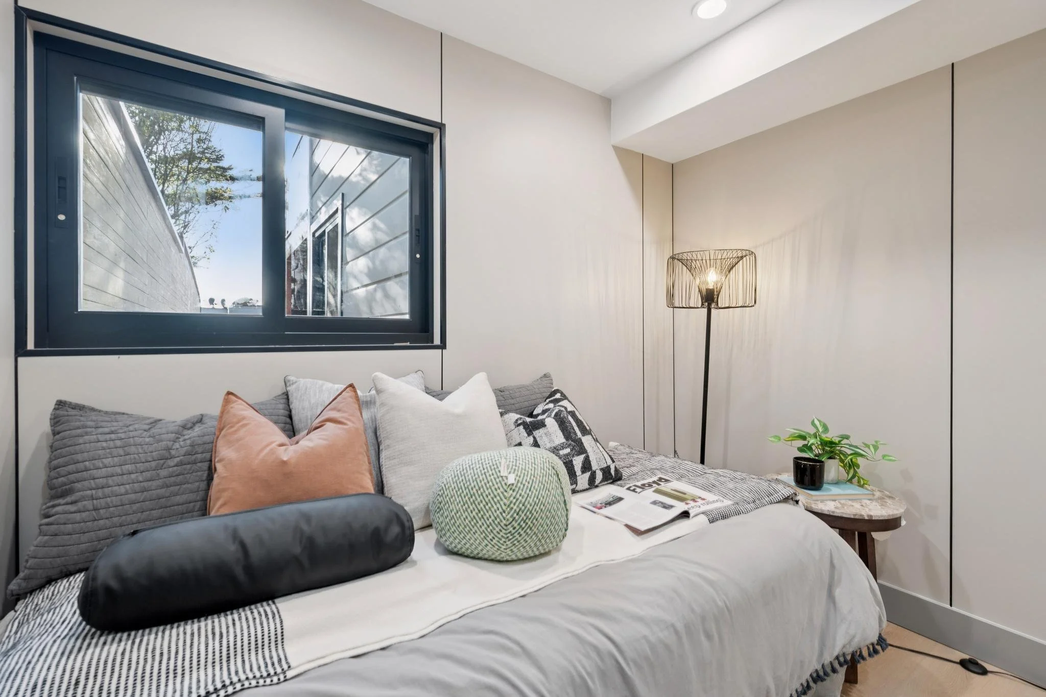 A cozy bedroom corner with a window, a bed with multiple pillows, a blanket, an open magazine, a potted plant, and a floor lamp in warm lighting.