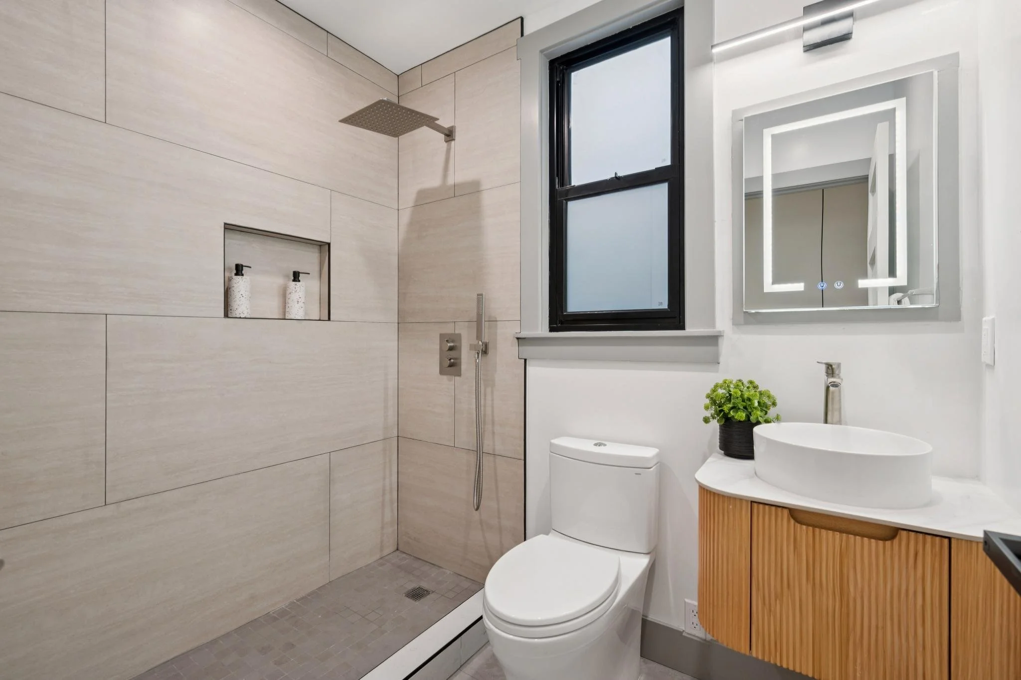 Modern bathroom with a walk-in shower, a toilet, a window, and a mirror with integrated lighting, featuring a wooden vanity with a vessel sink and a potted plant.