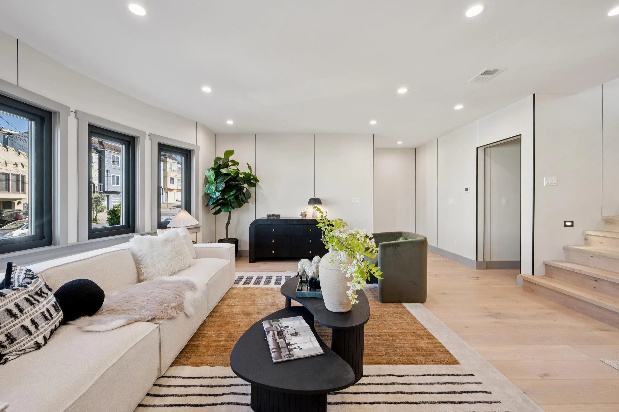 Living room with white sofa, black coffee table with decorative items, green armchair, large potted plant, window view of neighboring houses, coat rack, and stairs, modern decor, light wood flooring, white walls, ceiling lights.