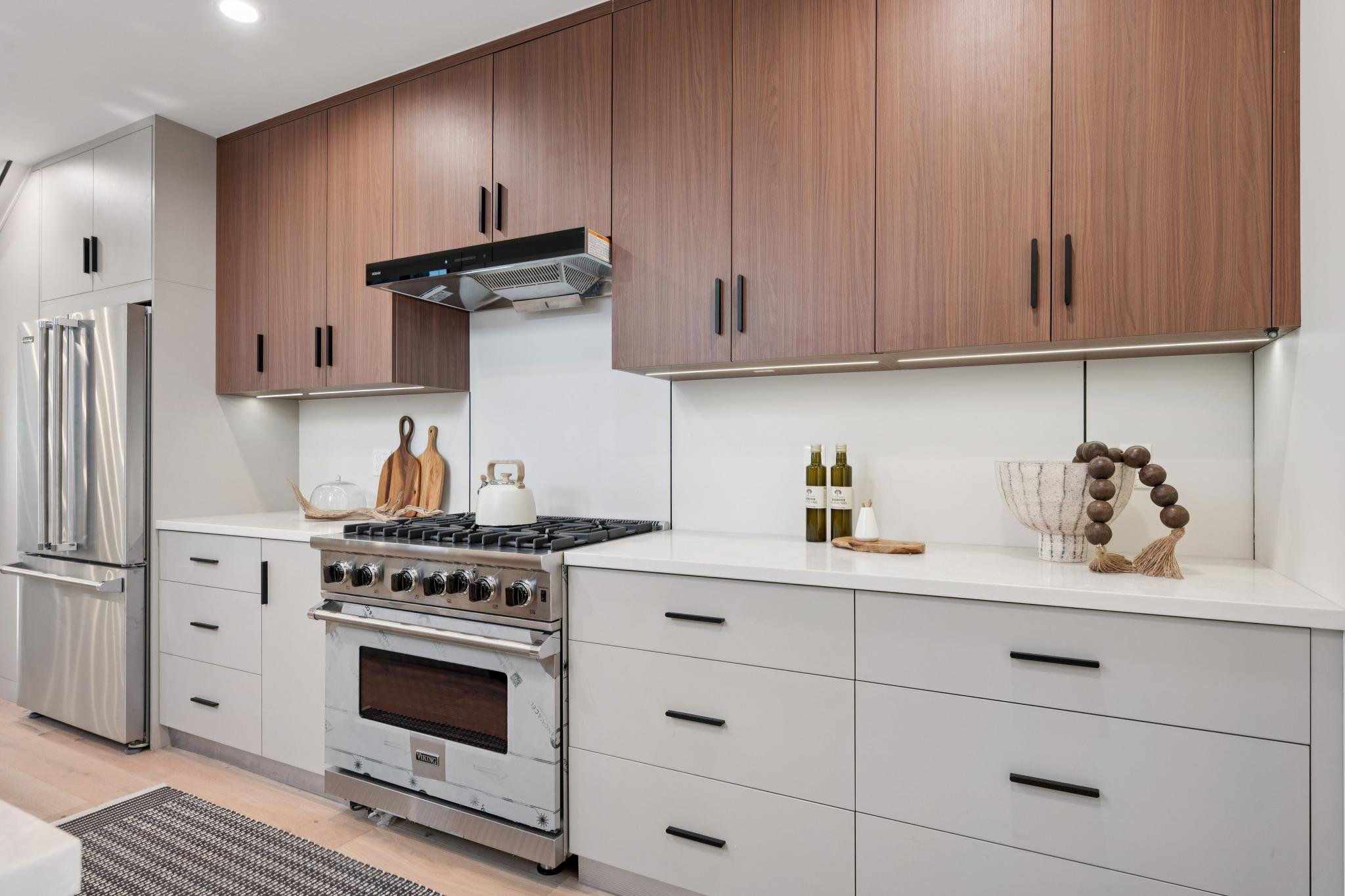 Modern kitchen with white lower cabinets, wood upper cabinets, stainless steel refrigerator and oven, white countertop, and decorative items including bottles, a bowl, and wooden cutting boards.