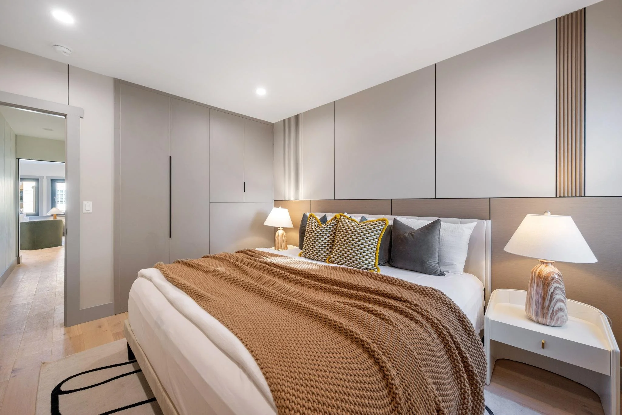 A neatly made bed with a tan knitted blanket, several decorative pillows, and white sheets, flanked by two bedside tables with lamps, in a modern, well-lit bedroom.