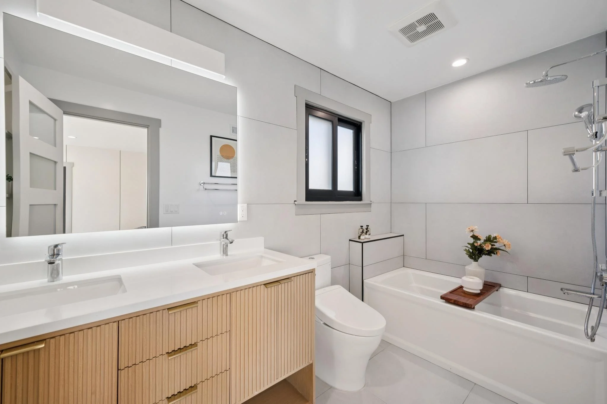 Modern bathroom with a double sink vanity, large mirror, toilet, bathtub with a wood tray, flower vase, window, and minimalistic decor.