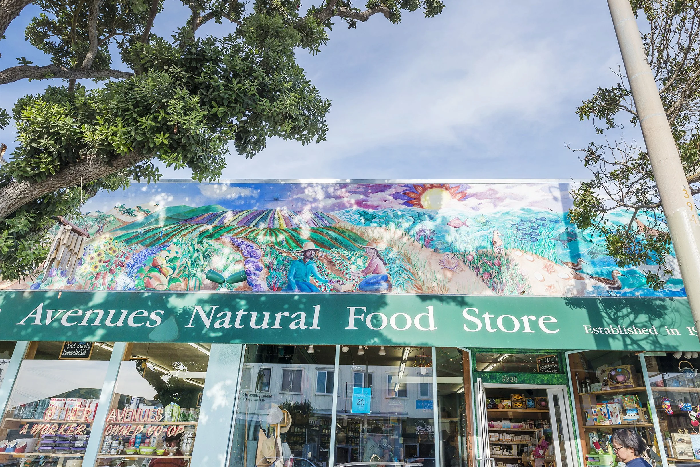 The storefront of Avenues Natural Food Store with a large colorful mural above the sign, showing a farm scene with people harvesting crops, sun in the sky, and aquatic life, and a woman wearing a hat near the entrance.