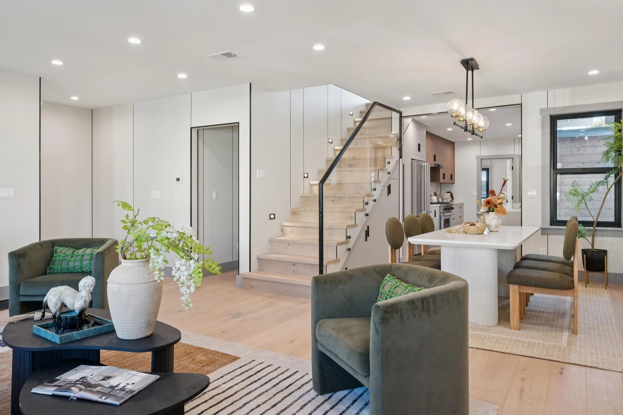 Modern open-concept living and dining area with light wood floors, green armchairs, a black coffee table with decorative items, and a white dining table with chairs, a staircase with glass railing, and kitchen area in the background.