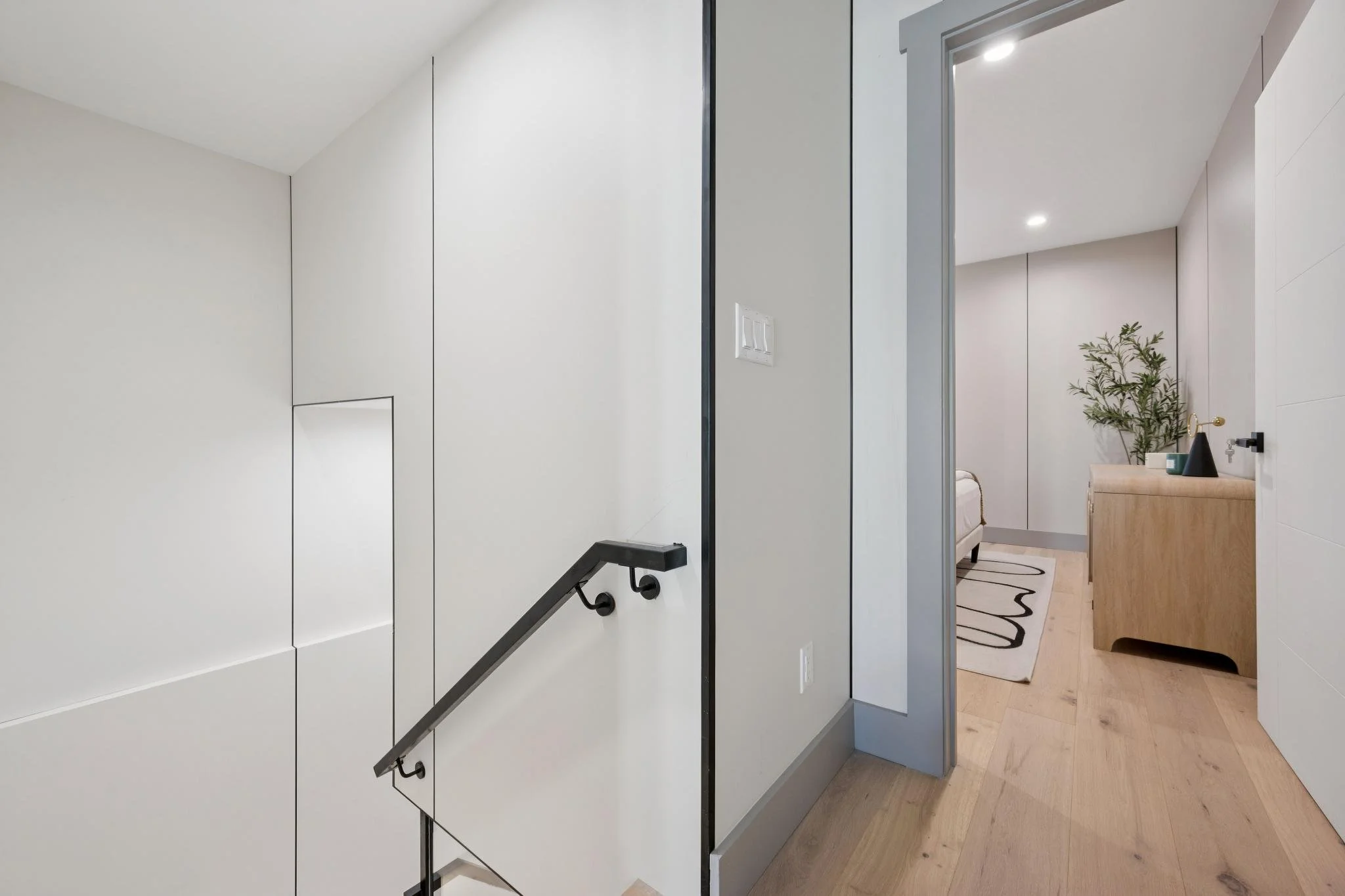 A modern interior hallway leading to a bedroom with beige walls, wooden flooring, and a small wooden desk with a decorative plant.