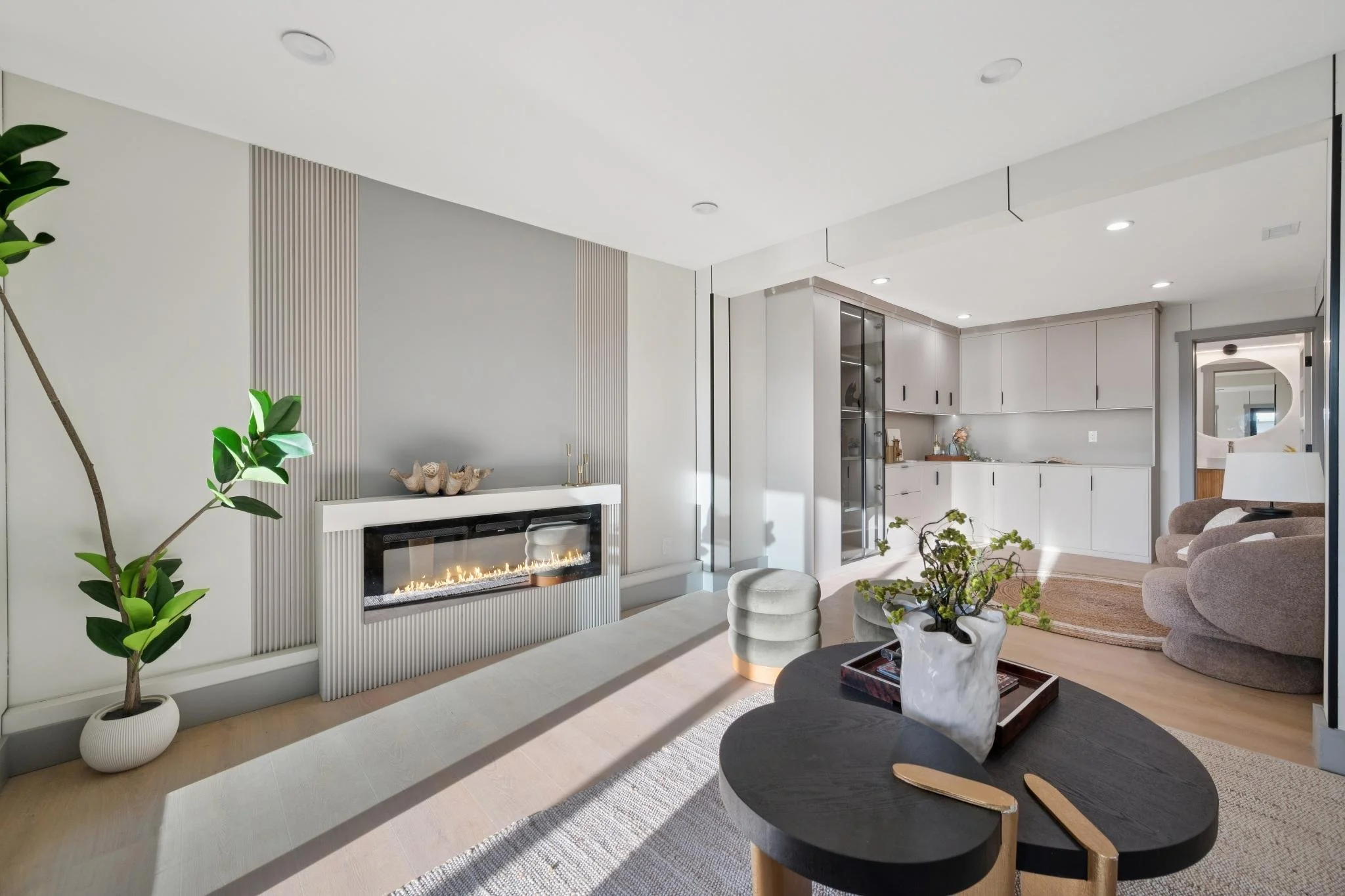 Modern living room with a built-in fireplace, large gray sectional sofa, round coffee table with a plant, and a kitchenette in the background.