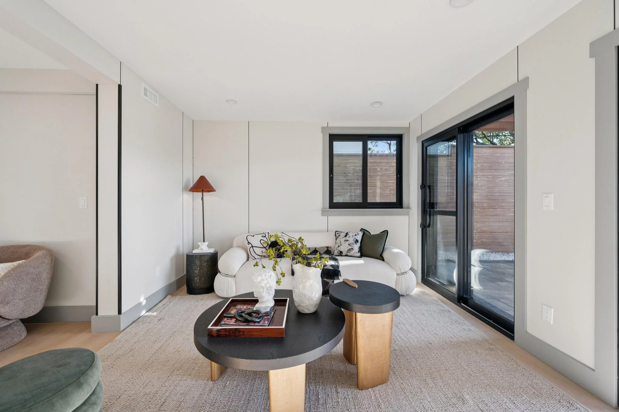 Living room with white sofa, black and white pillows, wooden coffee tables, a black side table, a floor lamp, windows, and a sliding glass door on a beige rug.