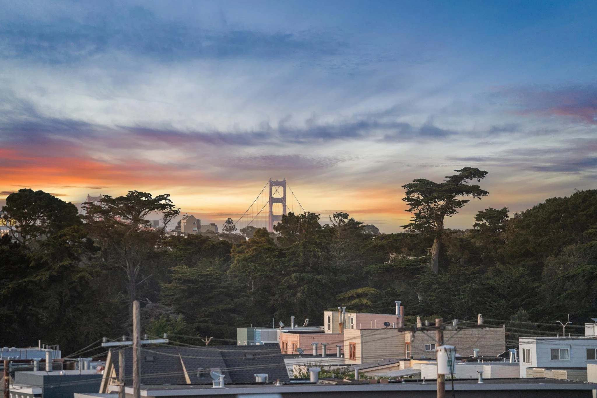 Cityscape at sunset with the Golden Gate Bridge visible in the background, trees in the foreground, and rooftops of residential buildings in the lower part of the image.
