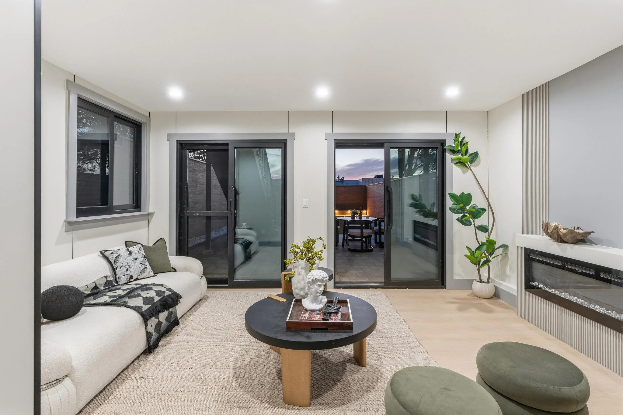 Living room with white sofa, black coffee table with sculptures and plants, sliding glass doors leading to balcony, and large window with black frames, decorated in neutral tones with modern decor and green plants.