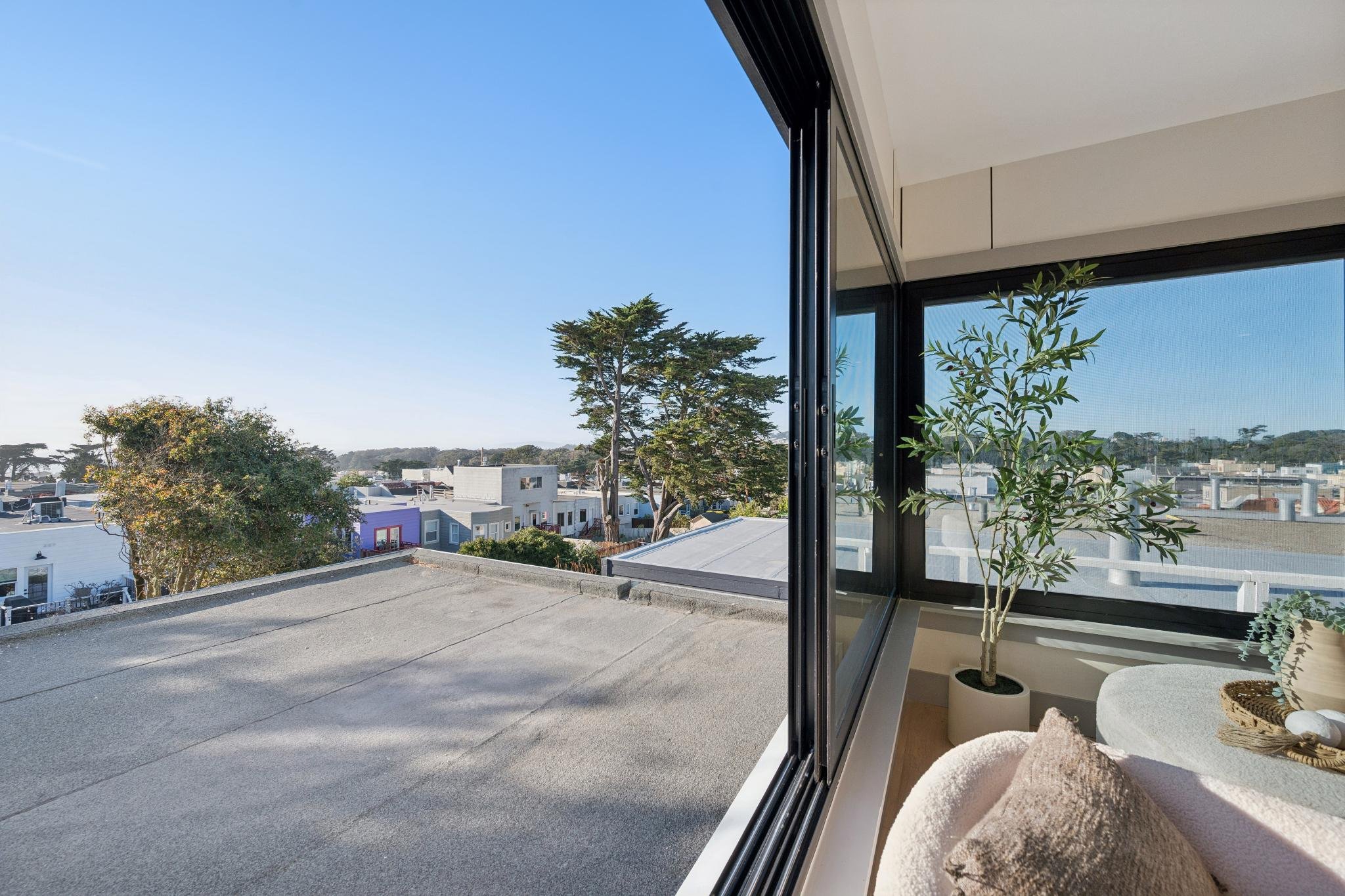 View from inside a room looking out onto rooftops and trees on a clear sunny day, with a large window and a potted plant inside.
