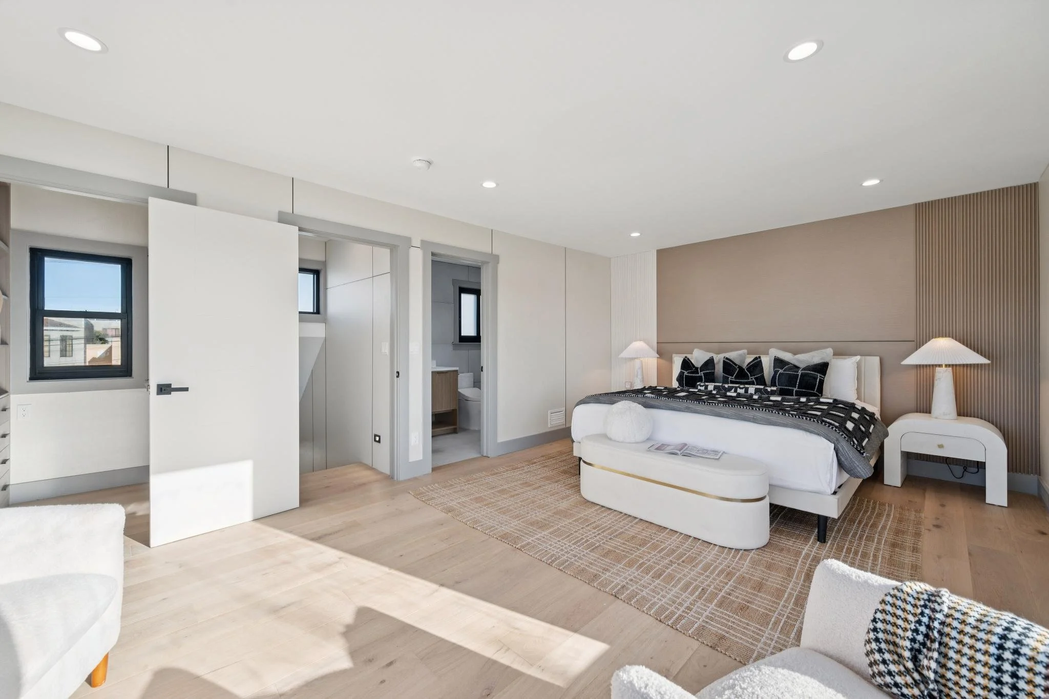 Modern bedroom with a white bed, black and white pillows, nightstands with lamps, and a small rug on light wood flooring. An open door reveals a bathroom and a closet with windows on the wall.