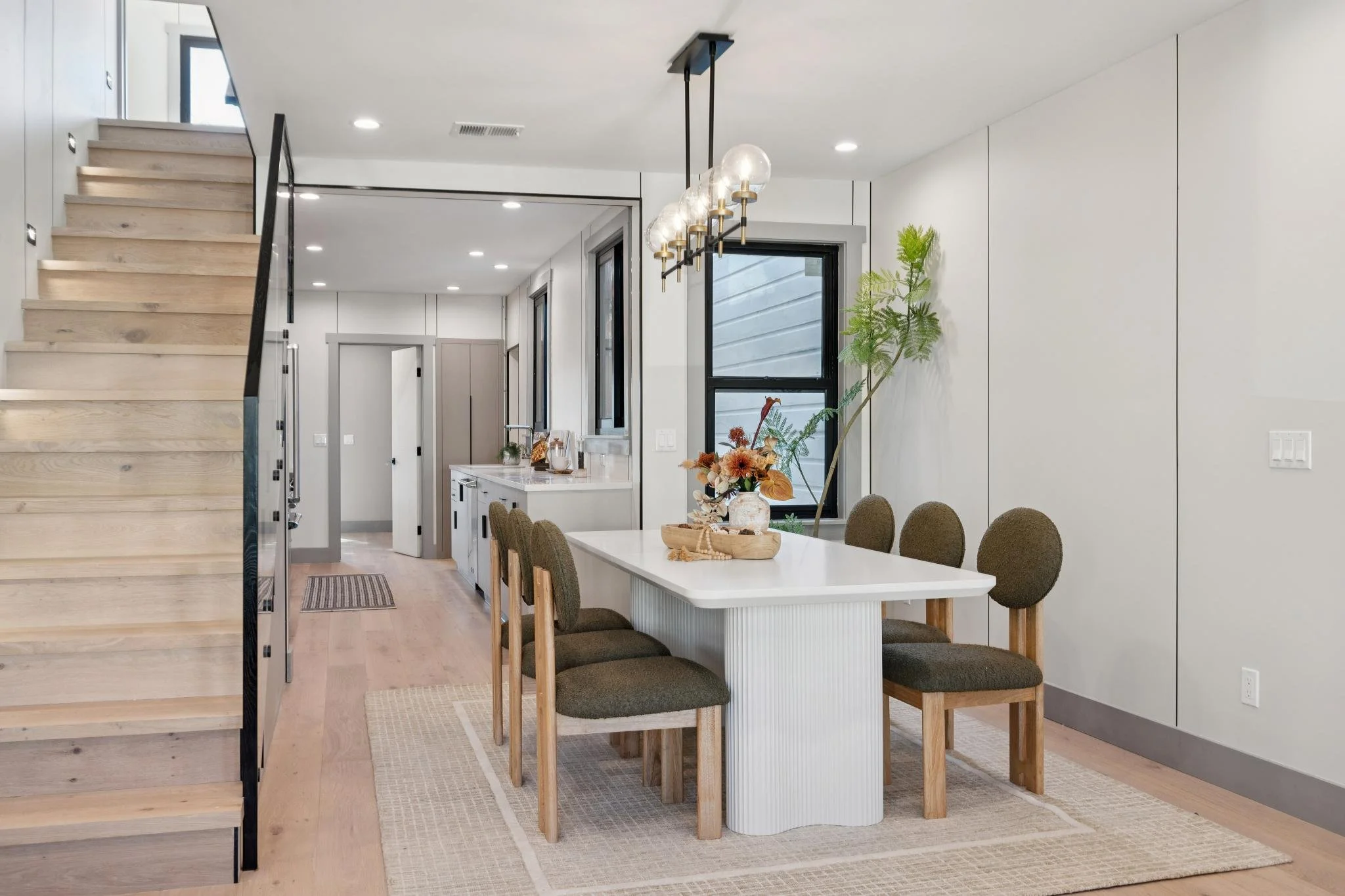 Dining area with white table, six brown chairs, wood flooring, staircase, and kitchen with white cabinets and black window frames.