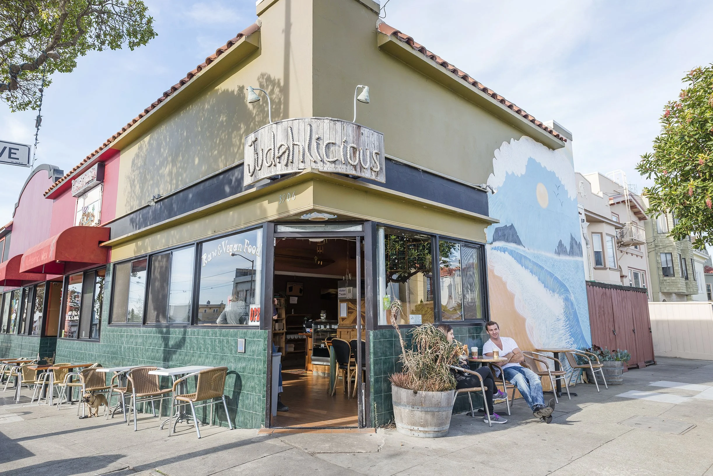 Exterior of a vegetarian and vegan restaurant called 'jiahlicious' with outdoor seating, a mural of a beach scene on the side of the building, and two people sitting outside enjoying drinks.