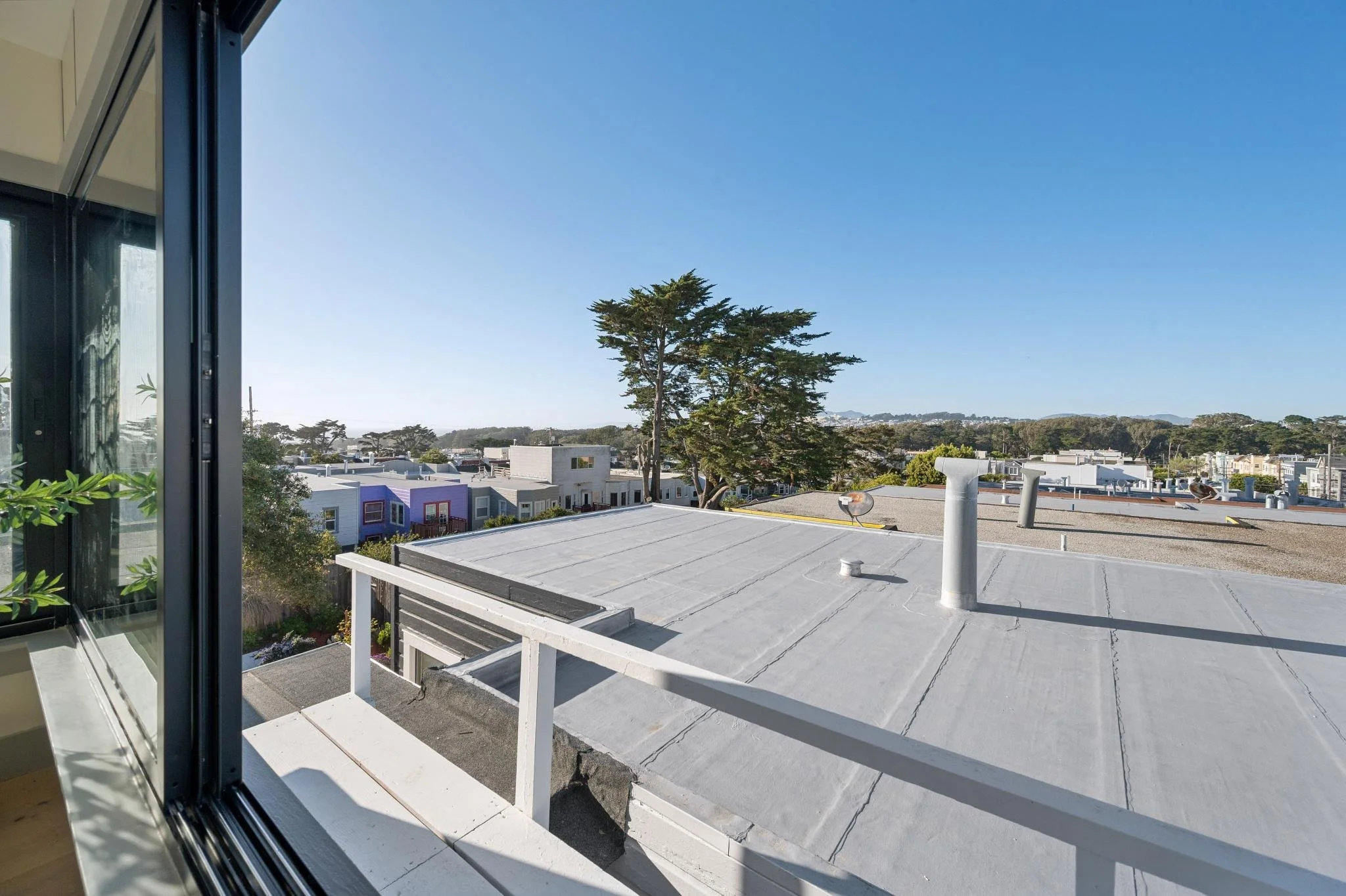 View from a window showing rooftops of residential buildings, trees, and a clear blue sky.