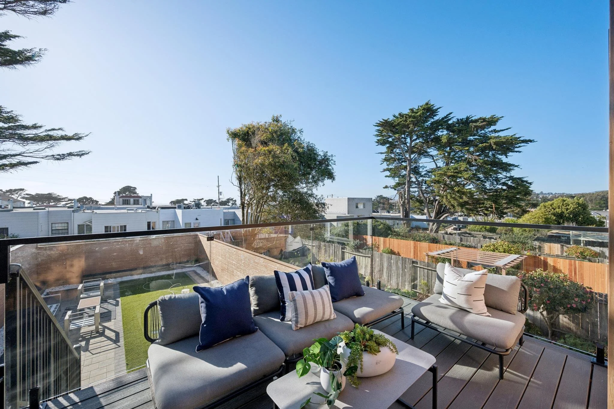 View of an outdoor balcony with seating, blue and white pillows, a white table with plants, overlooking neighboring houses and trees under a clear blue sky.