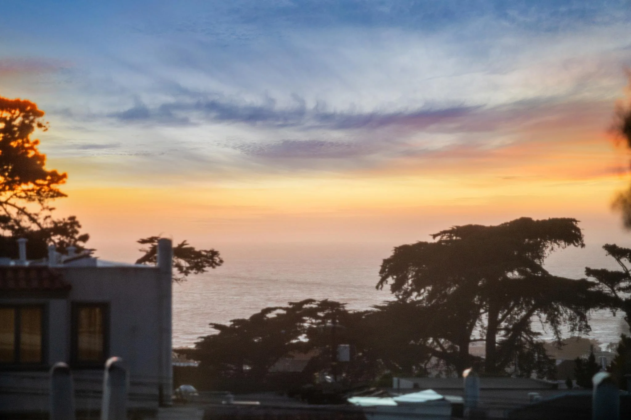 Sunset over the ocean with a colorful sky, silhouettes of trees and rooftops in the foreground.