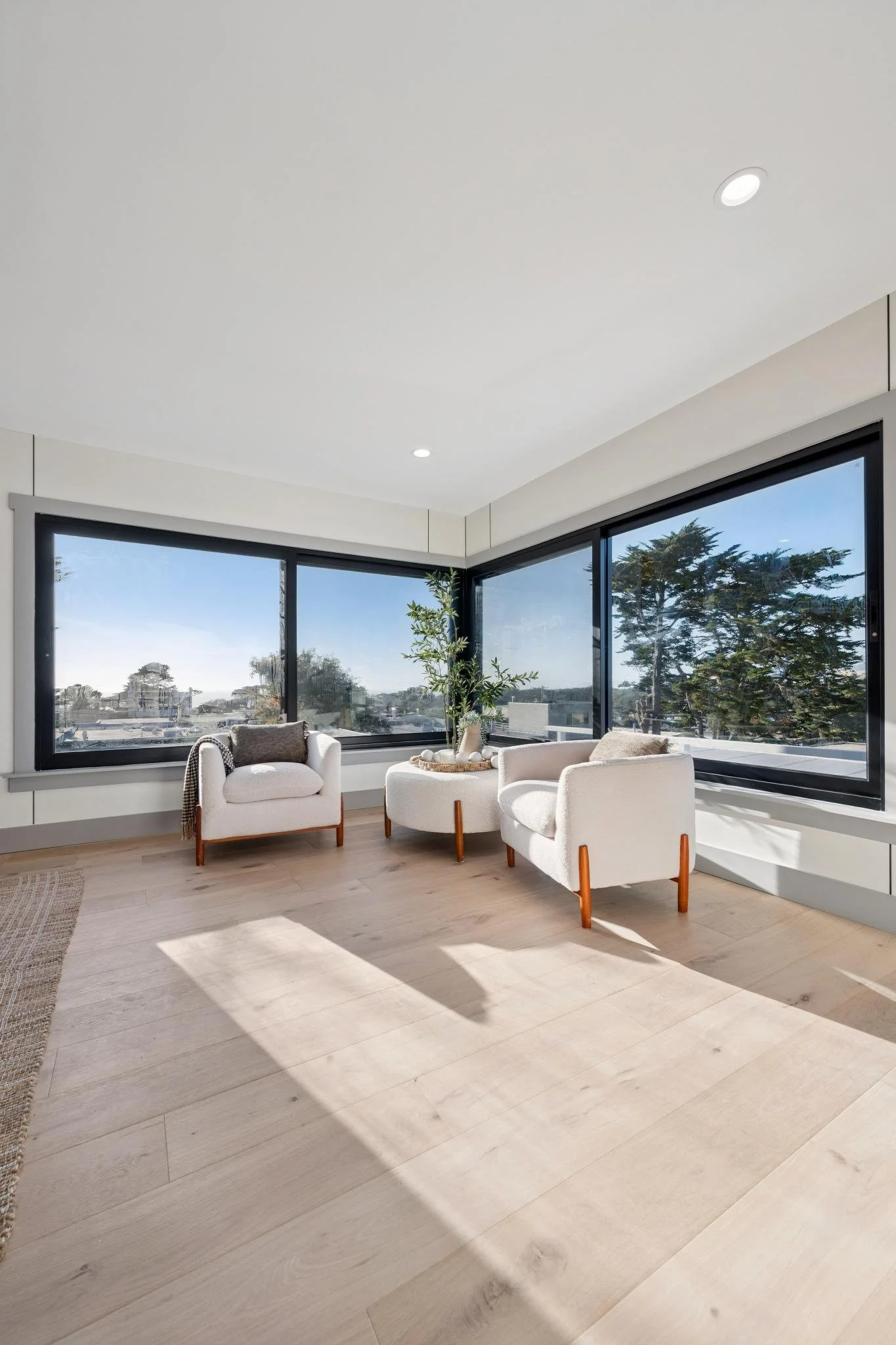 Empty room with large windows, white armchairs, a potted plant, and hardwood flooring, with sunlight casting shadows.