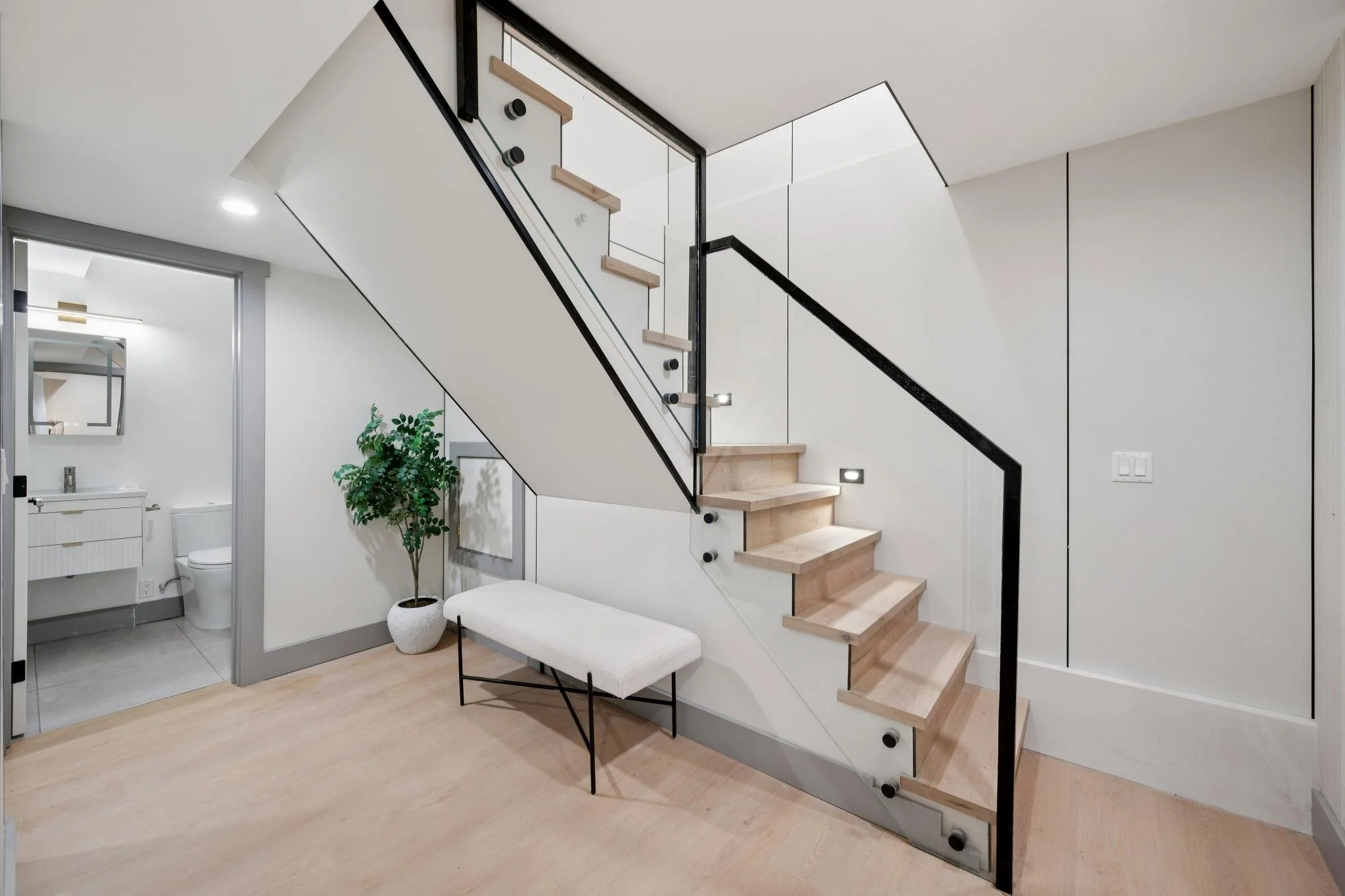 Modern staircase with light wooden steps and black metal railing in a minimalist interior with a small bench and potted plant, leading to upper floor, next to a white bathroom door.
