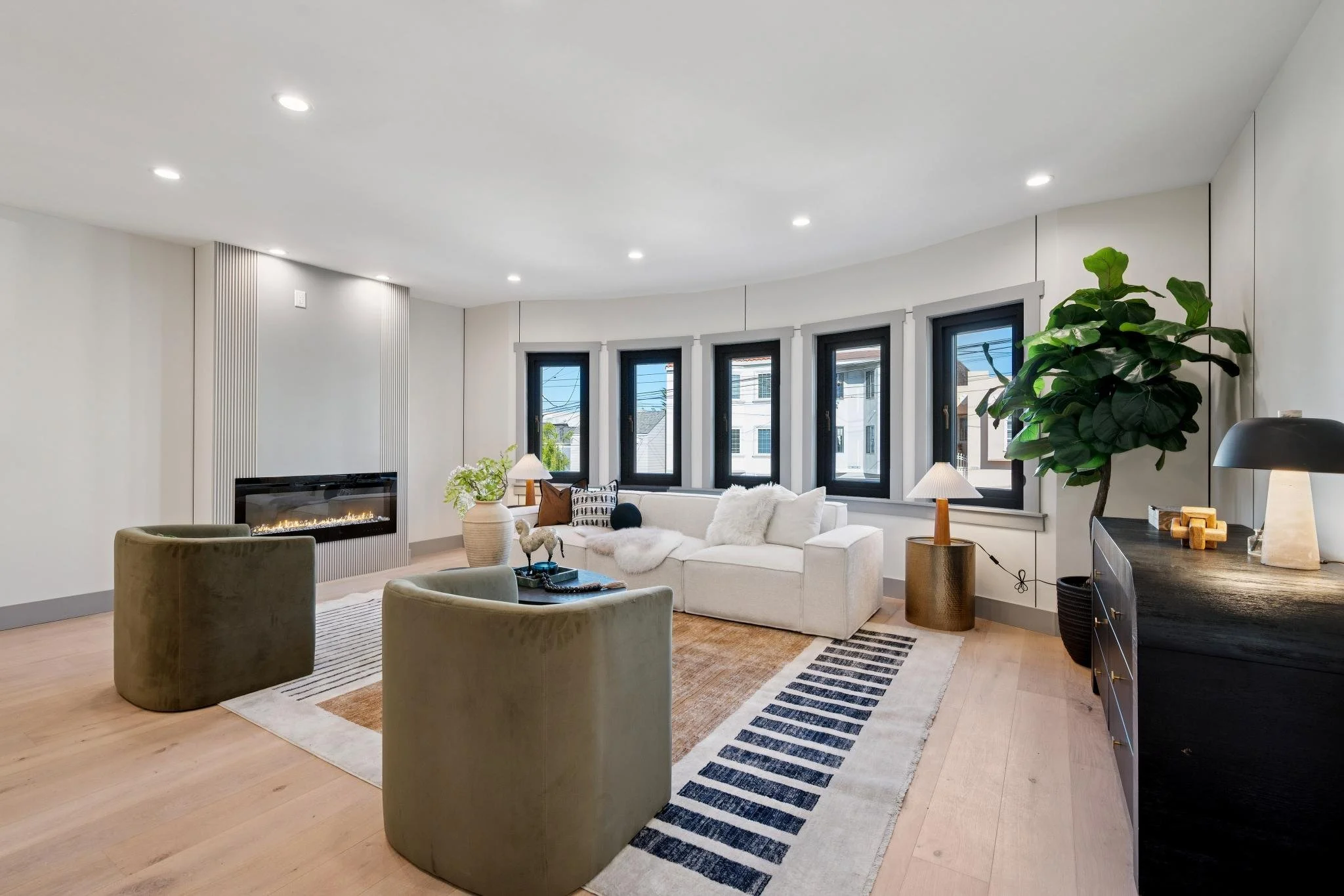 Modern living room with white sofa, green armchairs, a fireplace, black framed windows, and a large plant in a black pot.