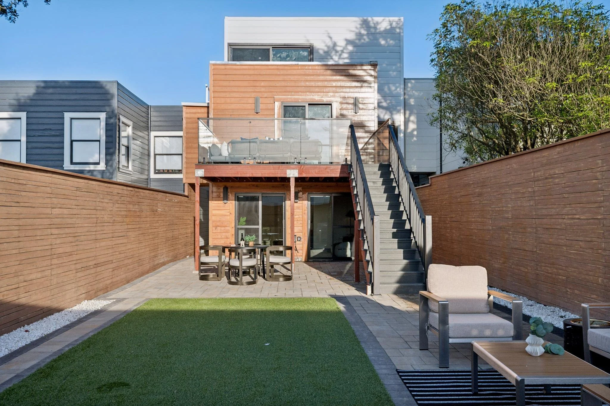 Backyard patio with outdoor seating, dining table, gray chairs, and an upstairs balcony with glass railing, surrounded by a tall wooden fence, on a sunny day with clear blue sky.