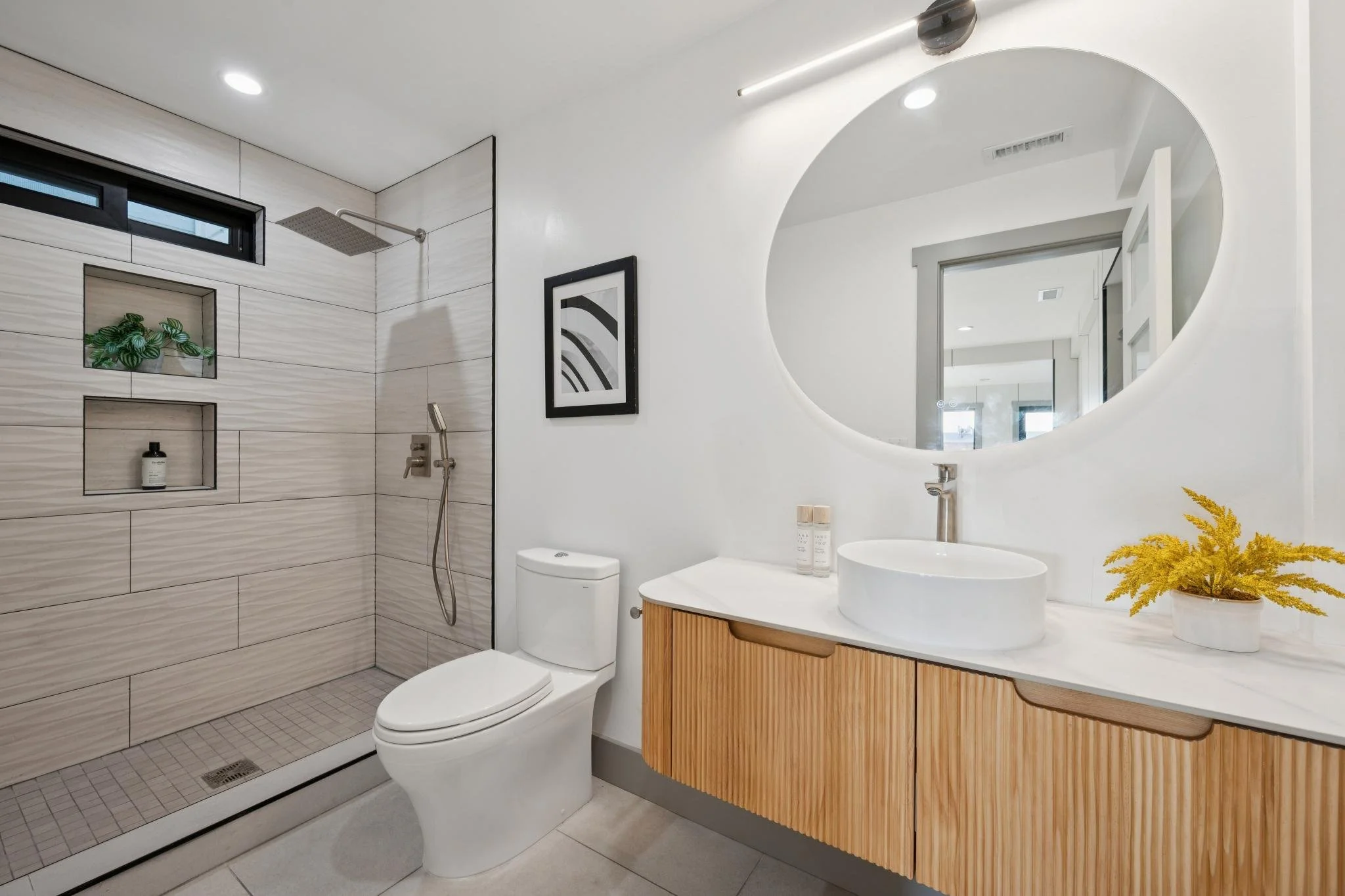 Modern bathroom with walk-in shower, toilet, and vanity with round vessel sink, large oval mirror, framed black-and-white abstract art, and yellow decorative plant.