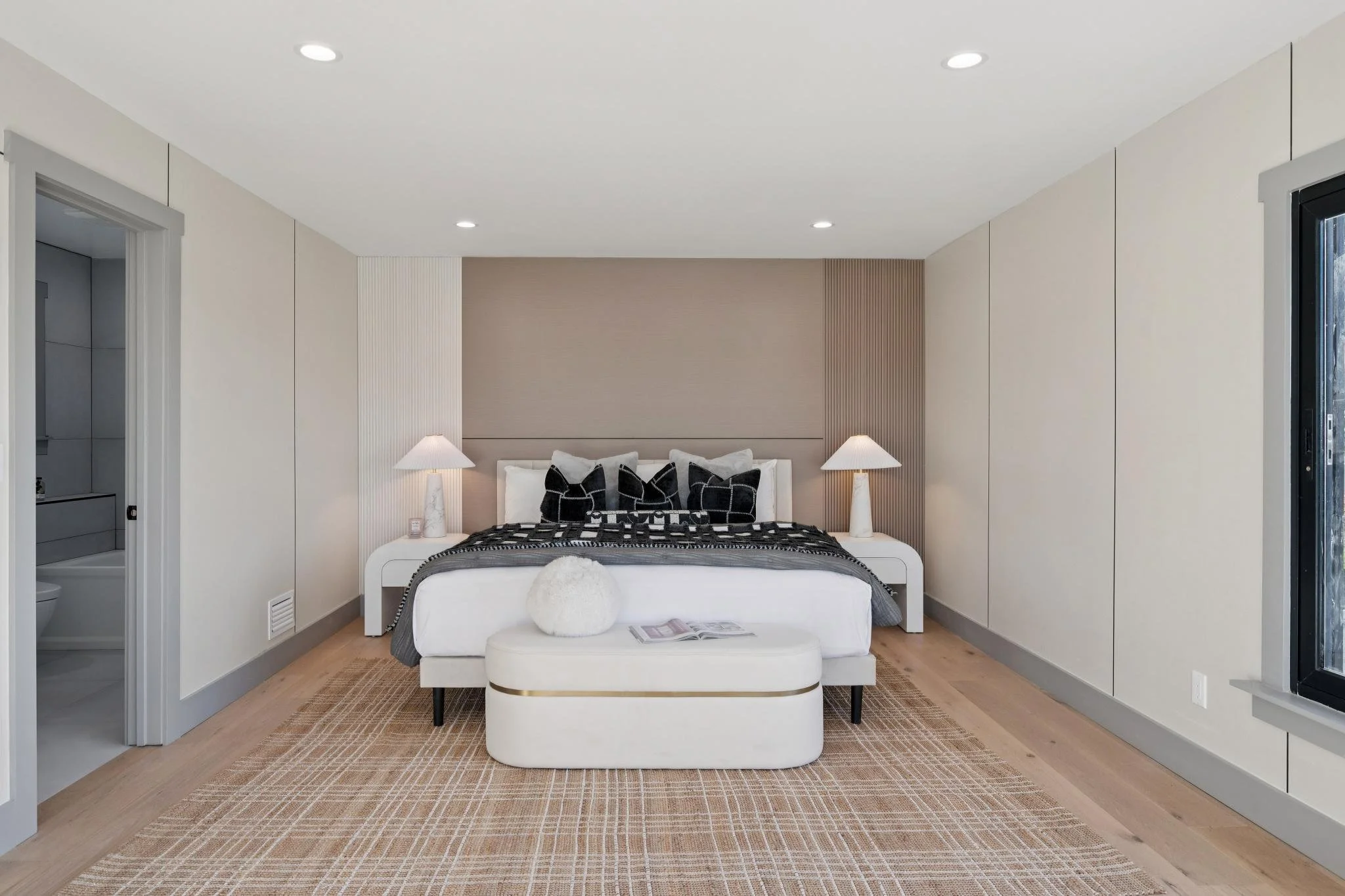 Modern bedroom with a white bed, black and white pillows, and two matching lamps on white nightstands. A plush bench with a book and decorative pillow is at the foot of the bed, on a beige woven rug. The room features minimalist decor with beige wall