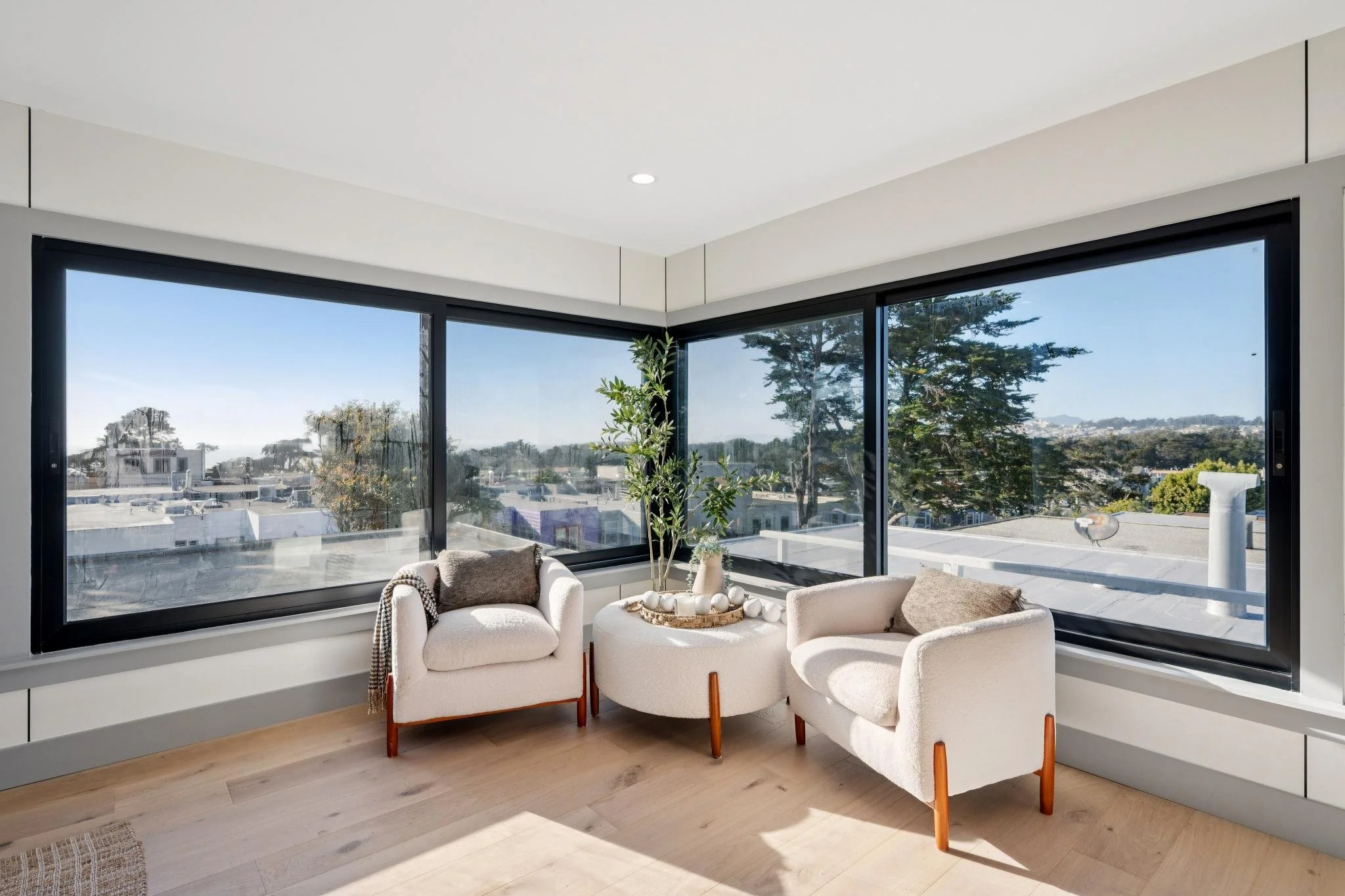 Living room with large corner windows, two cream-colored armchairs, a small round table with decorative white vases, and a tall green plant against a view of rooftops and trees outside.