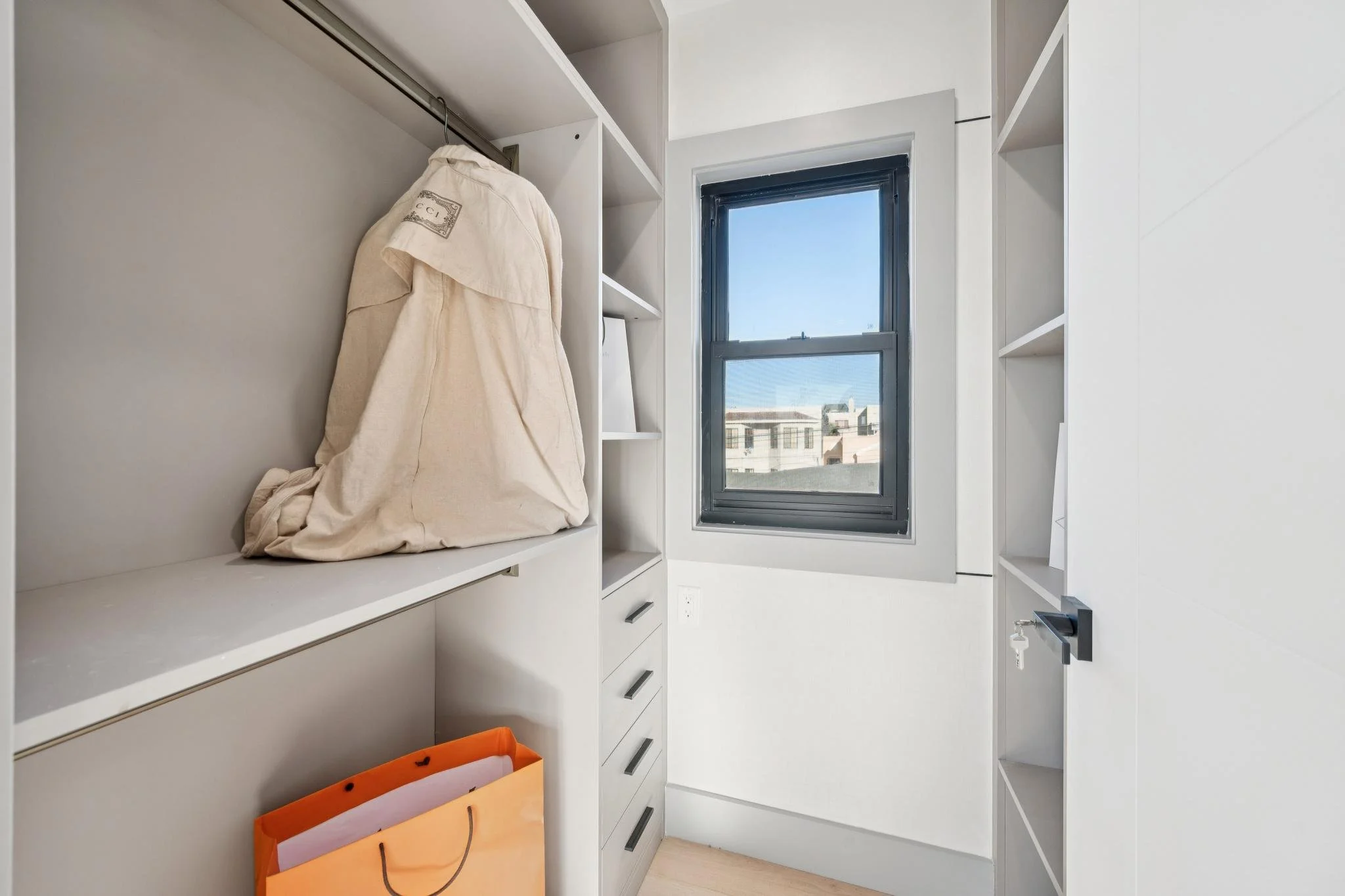 Empty walk-in closet with white shelves, a hanging rail with a beige fabric bag, a window with a view of buildings outside, and an orange shopping bag on the floor.