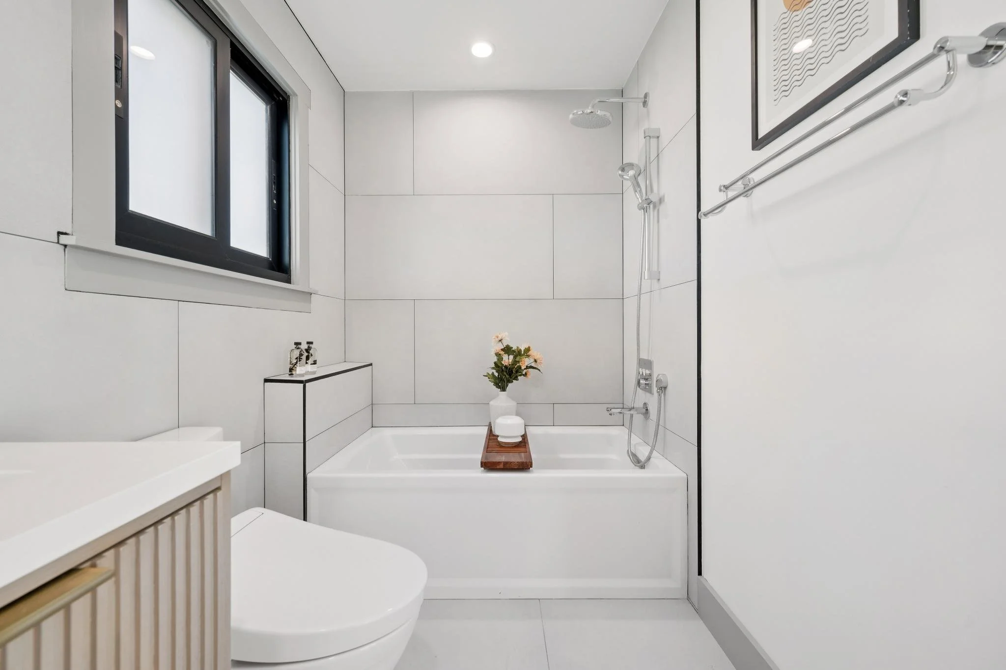 Minimalist bathroom with white large tiles, a bathtub with a wooden tray holding flowers and bath essentials, a window, and modern fixtures.