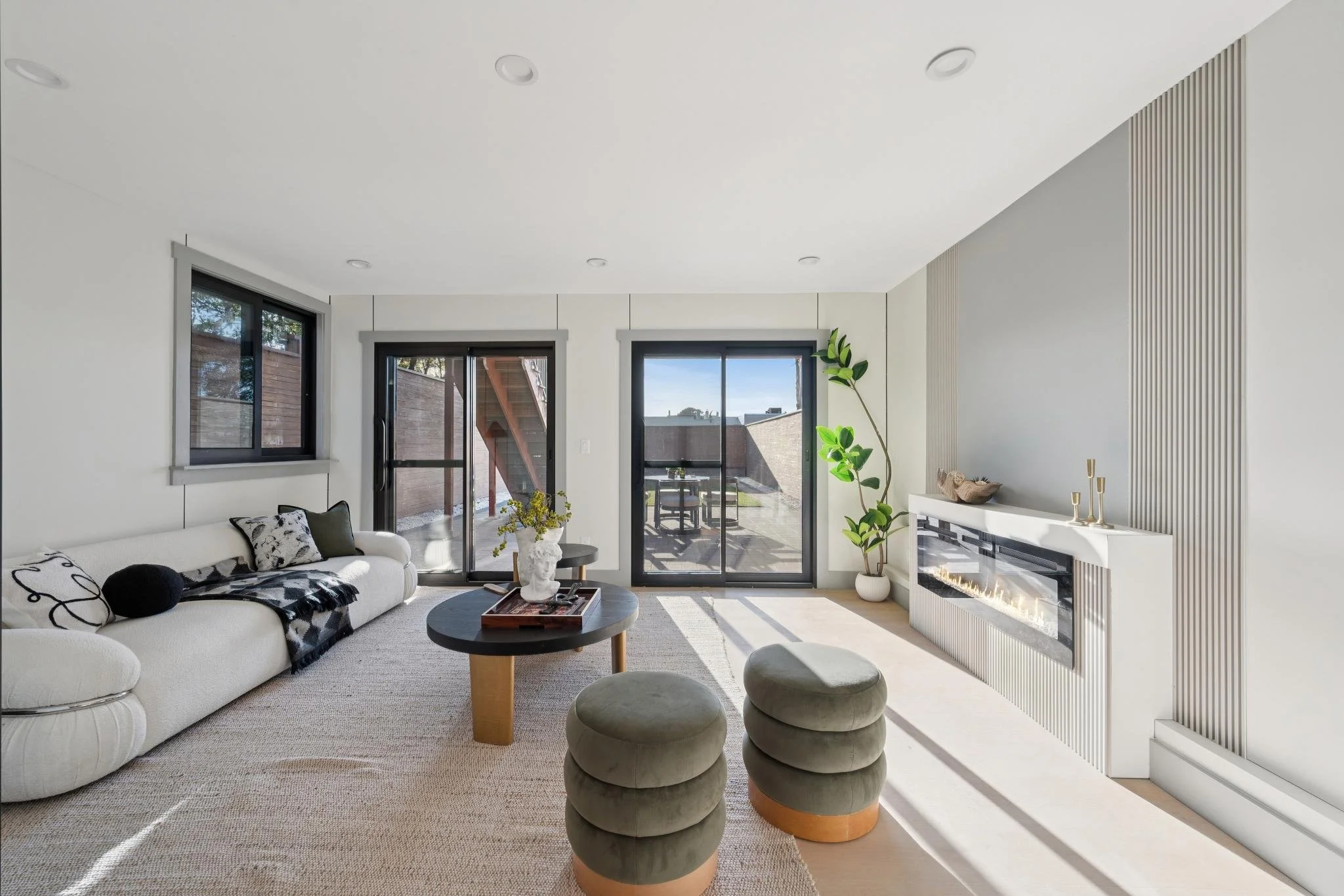 Modern living room with white sofa, black and white pillows, black coffee table, green plants, and a fireplace, with sliding glass doors leading outside.
