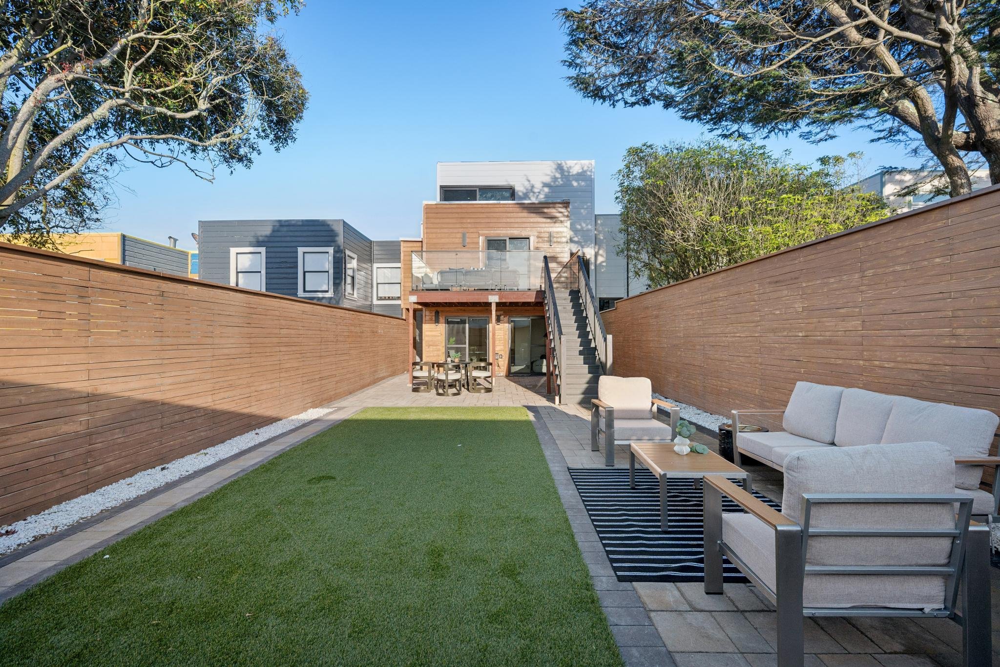Backyard with grass lawn, patio furniture, wooden fencing, and modern multi-story house in the background under a clear blue sky.