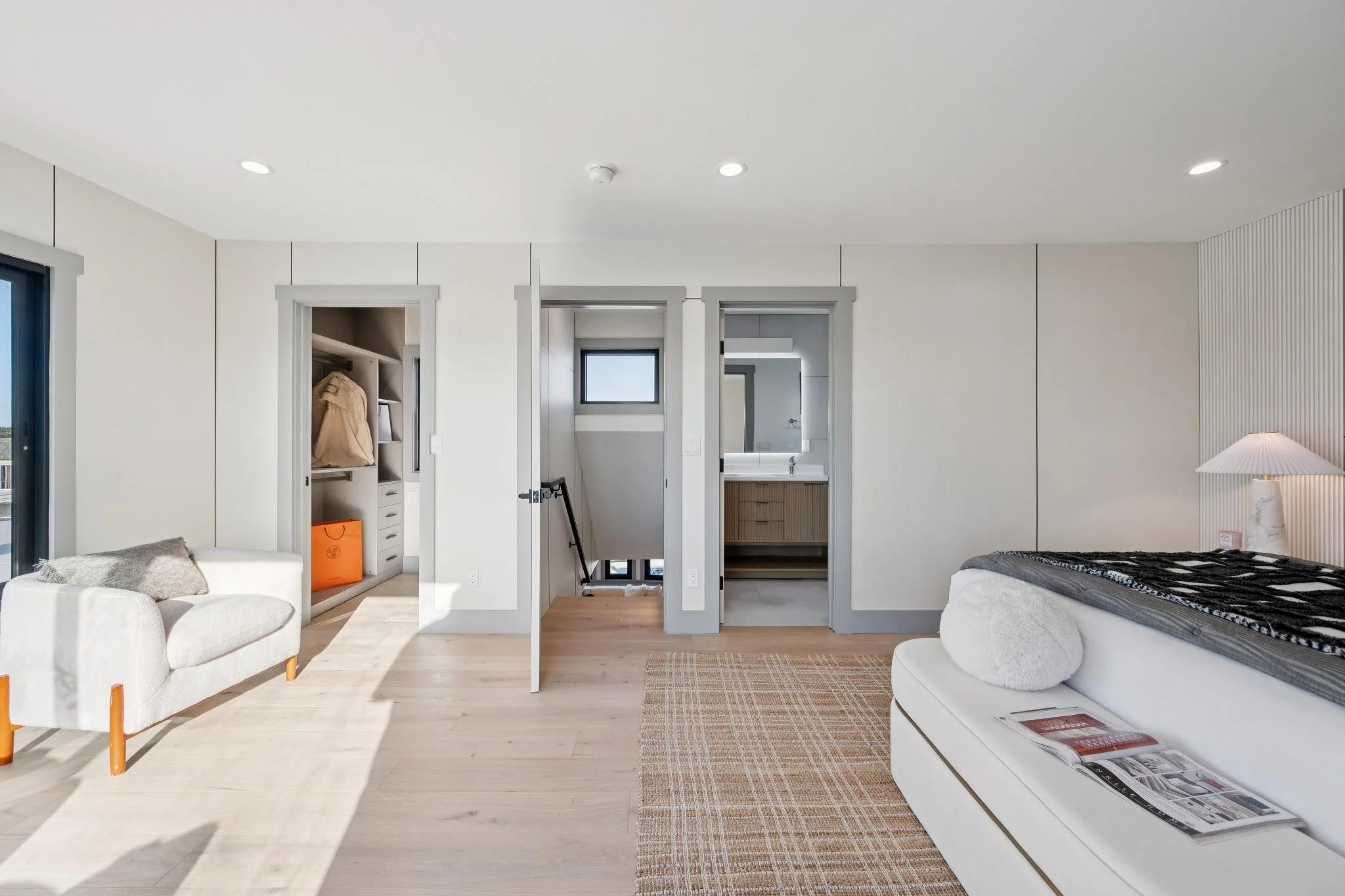 Modern bedroom with white walls, light wood flooring, a white couch with a fur pillow, a bed with a blanket and a magazine, a side table with a lamp, an open closet, and a bathroom with a vanity and mirror.