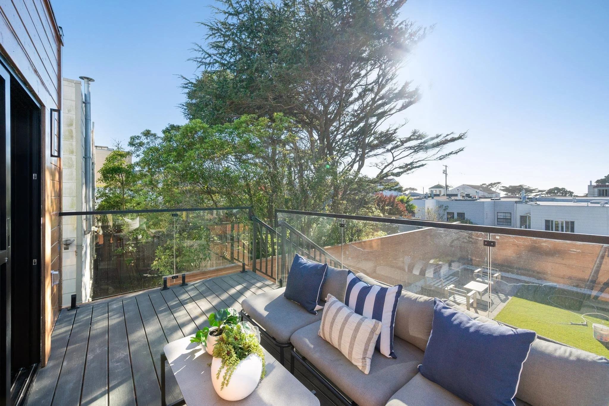 A balcony with a cushioned outdoor sofa decorated with various pillows, a white coffee table with a potted plant, surrounded by a glass railing, overlooking a yard and neighboring buildings with trees and clear blue sky.