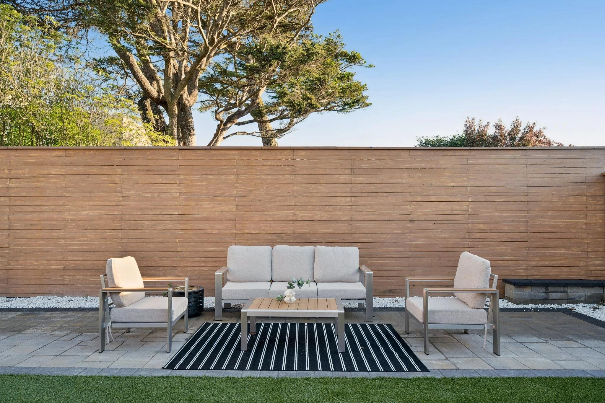 Outdoor patio with a cream-colored sofa and two matching armchairs arranged around a small rectangular table on a black and white striped rug. There are white stones along a wooden privacy fence and a large tree in the background with a clear blue sk