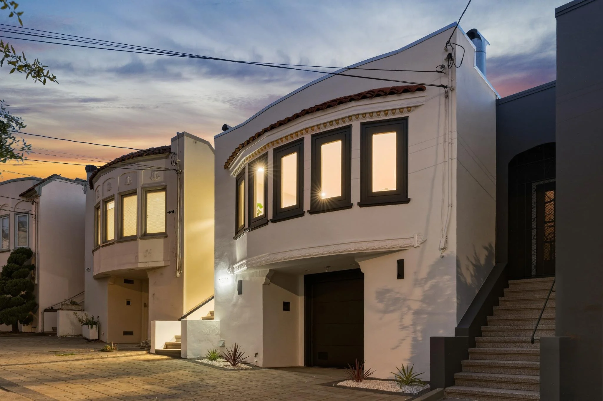 A row of modern white houses at dusk with illuminated windows and a sunset sky in the background.