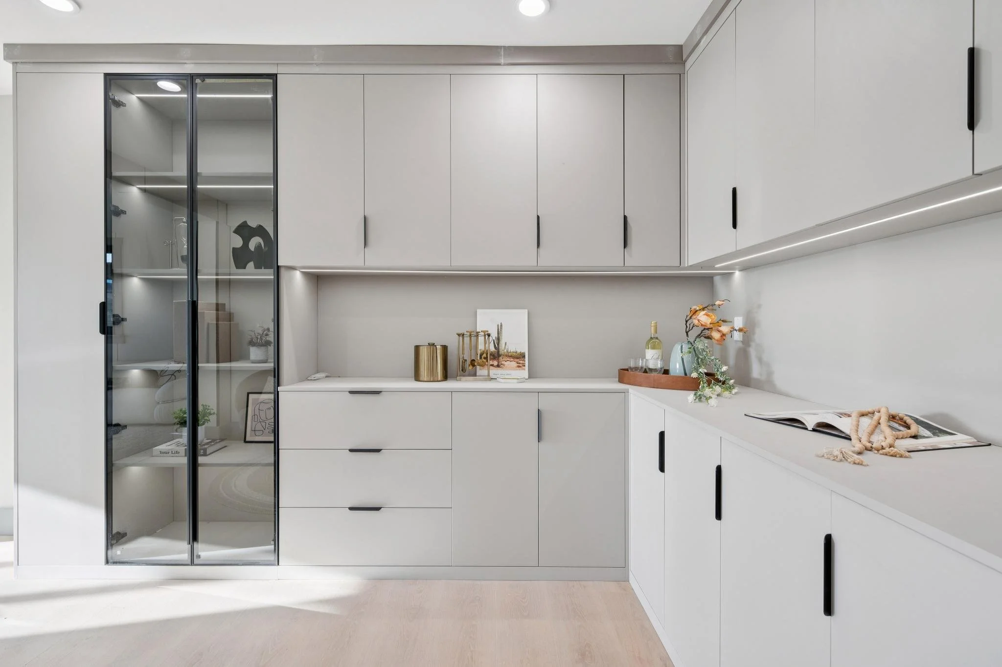 Modern white kitchen with black handles, corner countertop with decorative items, and glass-front cabinets with shelves and lighting.