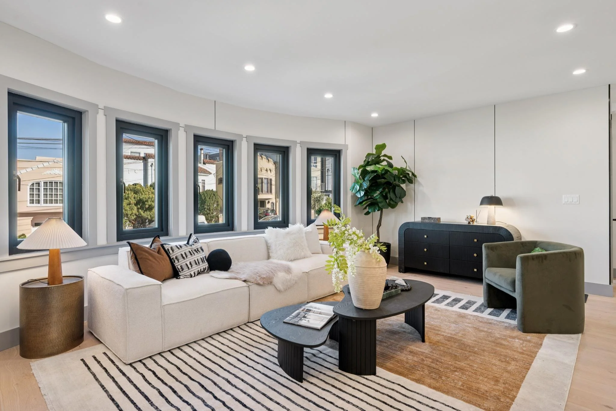 Modern living room with a white sofa, decorative pillows, a coffee table with a vase, large windows, and a sideboard, featuring minimalist decor and a potted plant.