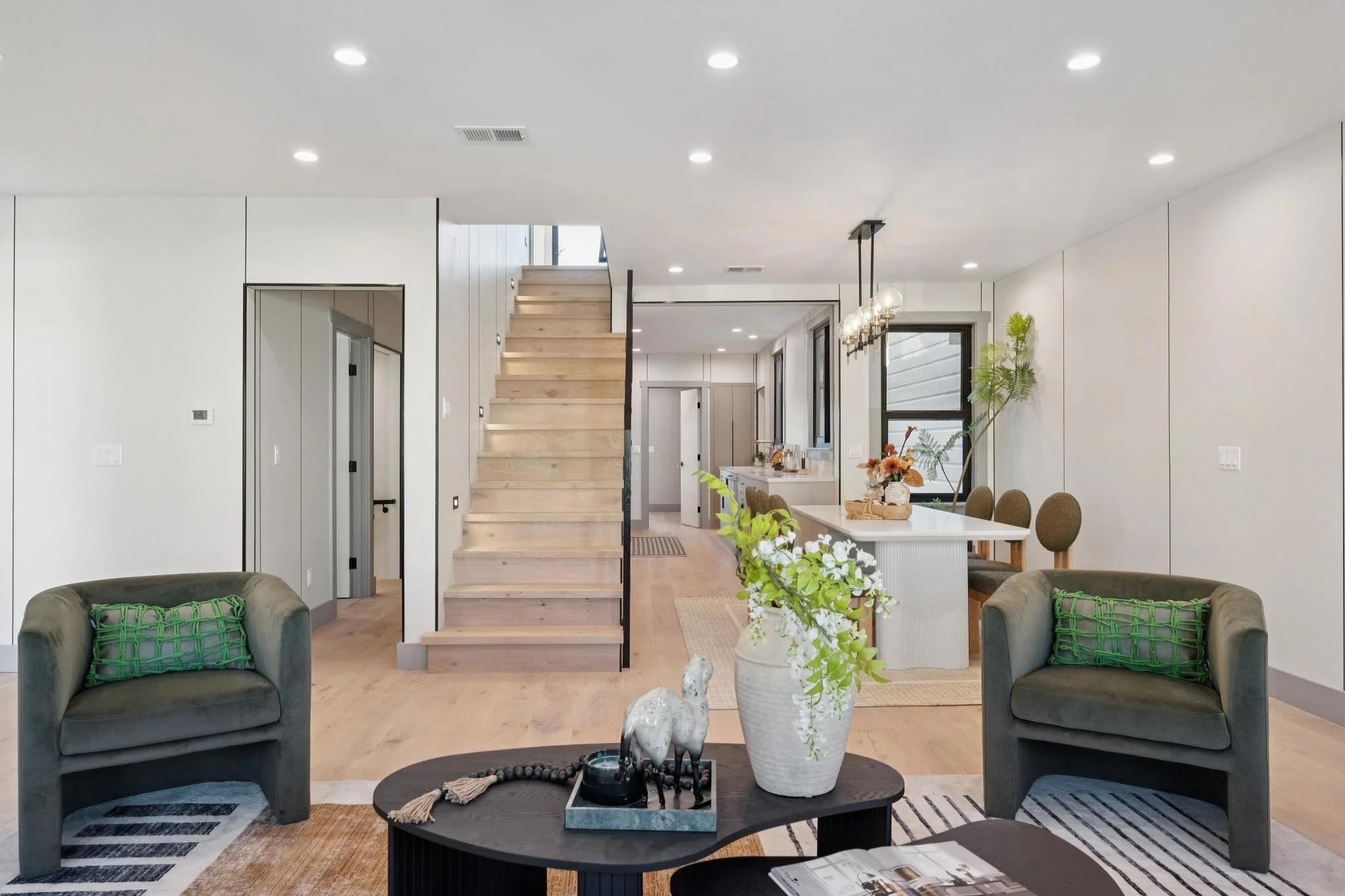 Modern living room with beige chairs, a round black coffee table with horse and llama figurines, and a white vase with floral arrangement. Open kitchen and staircase in the background.