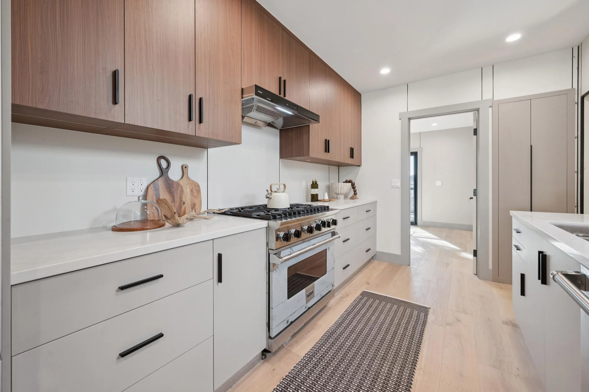 Modern kitchen with white cabinets, black handles, wooden upper cabinets, stainless steel oven, white countertop, decorative cutting boards, kettle, bowl, and a striped rug on wooden floor.