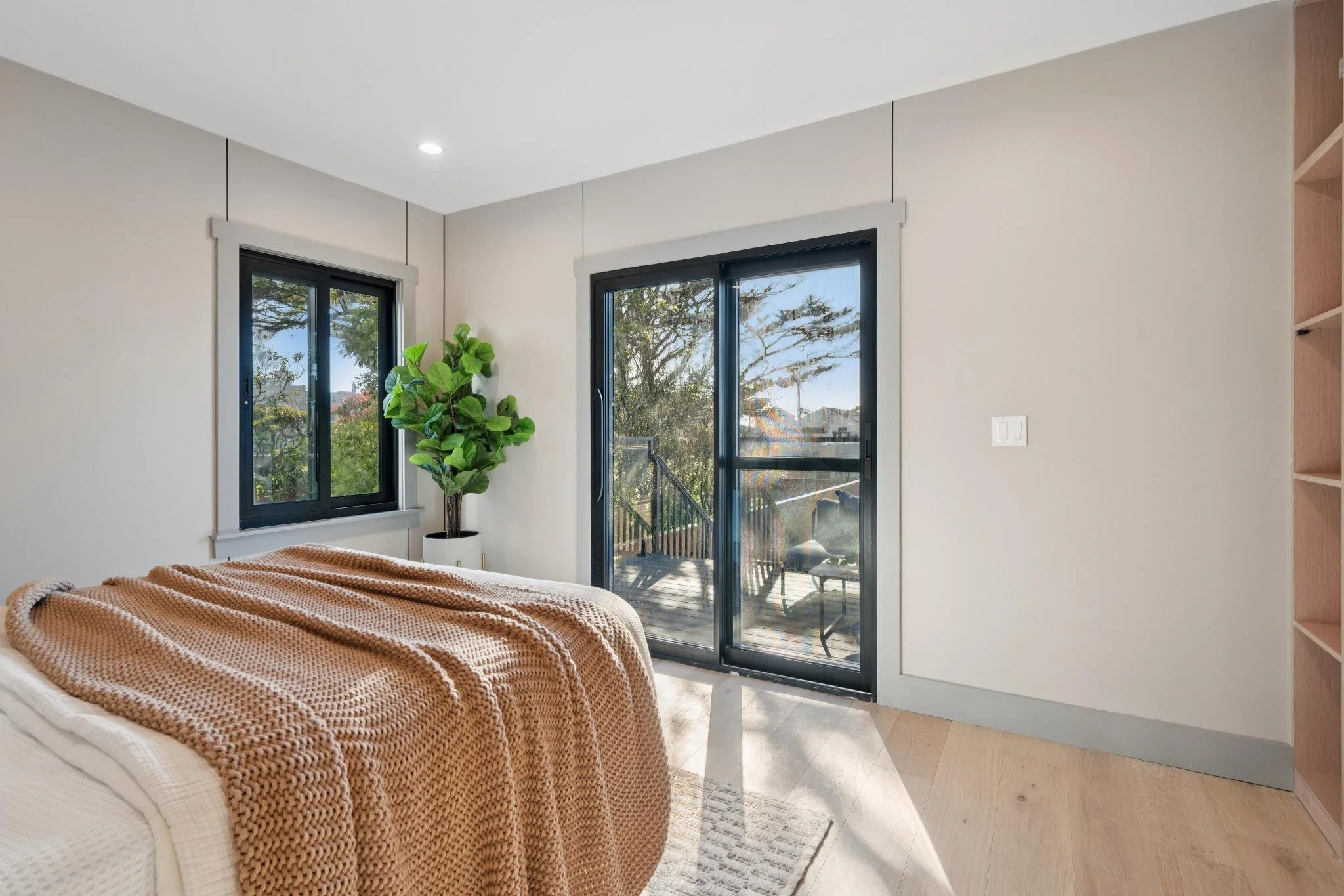 A bedroom with a bed covered in a beige knitted blanket, a green potted plant near a window, and sliding glass doors leading to a balcony with outdoor furniture, sunlit and with trees visible outside.