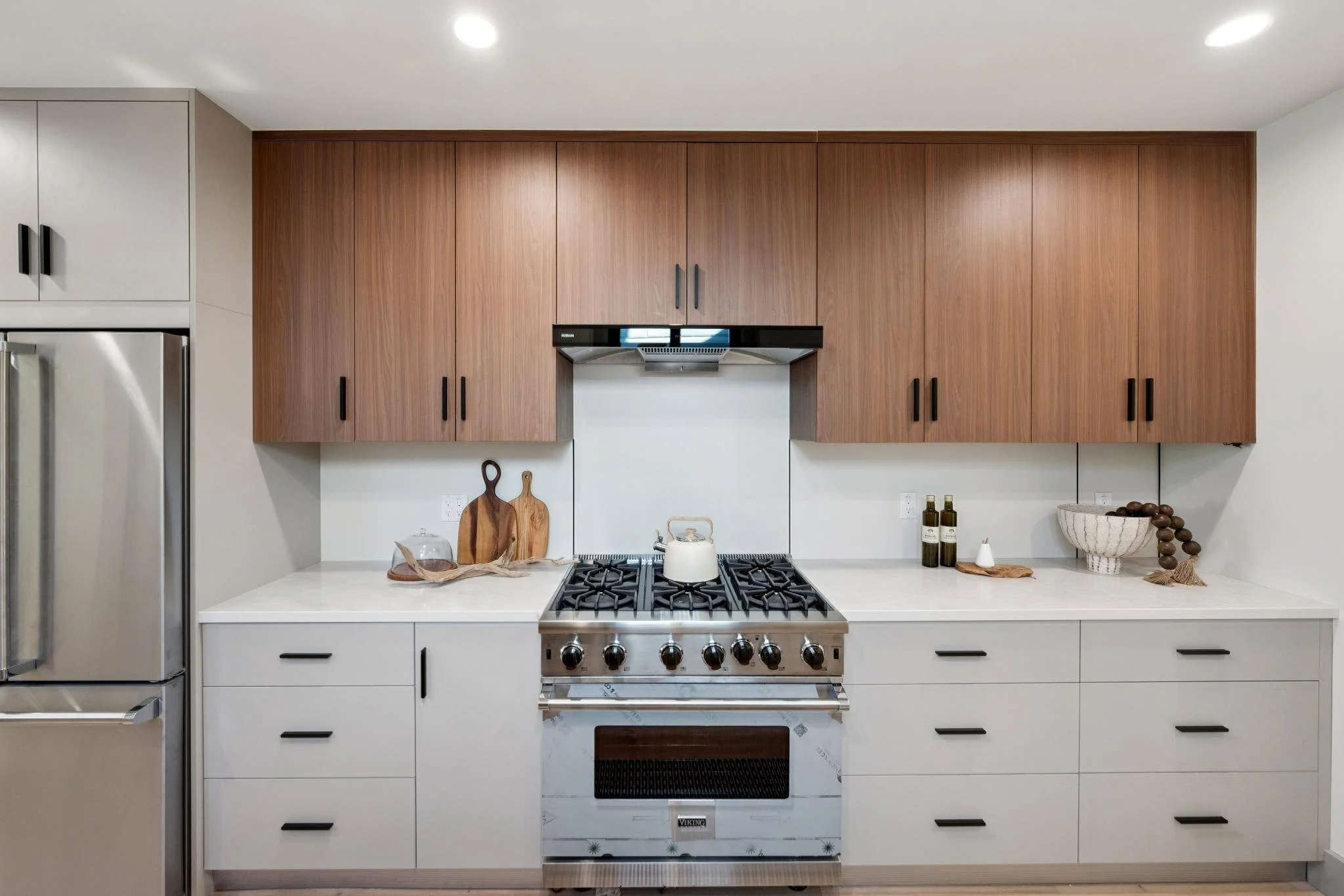 Modern kitchen with white cabinets, wooden upper cabinets, stainless steel stove with a tea kettle, and minimalist decor including cutting boards, bottles, and a bowl.